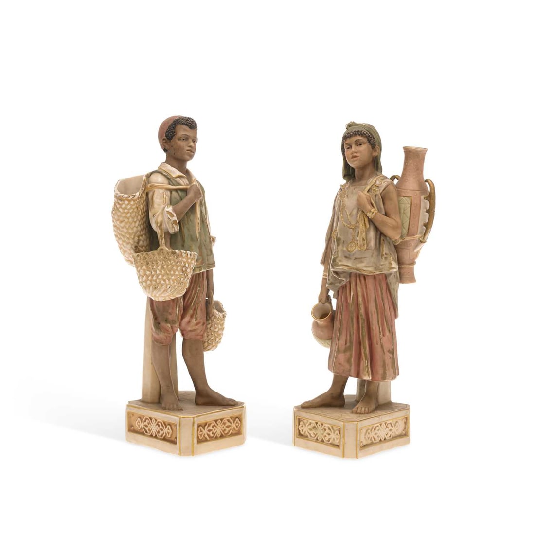 A PAIR OF ROYAL DUX FIGURES CIRCA 1910: A PAIR OF ROYAL DUX FIGURES CIRCA 1910 the figures well modelled as young North African children, carrying their wares to market, set upon canted square bases, Royal Dux, Bohemia triangle mark to the 