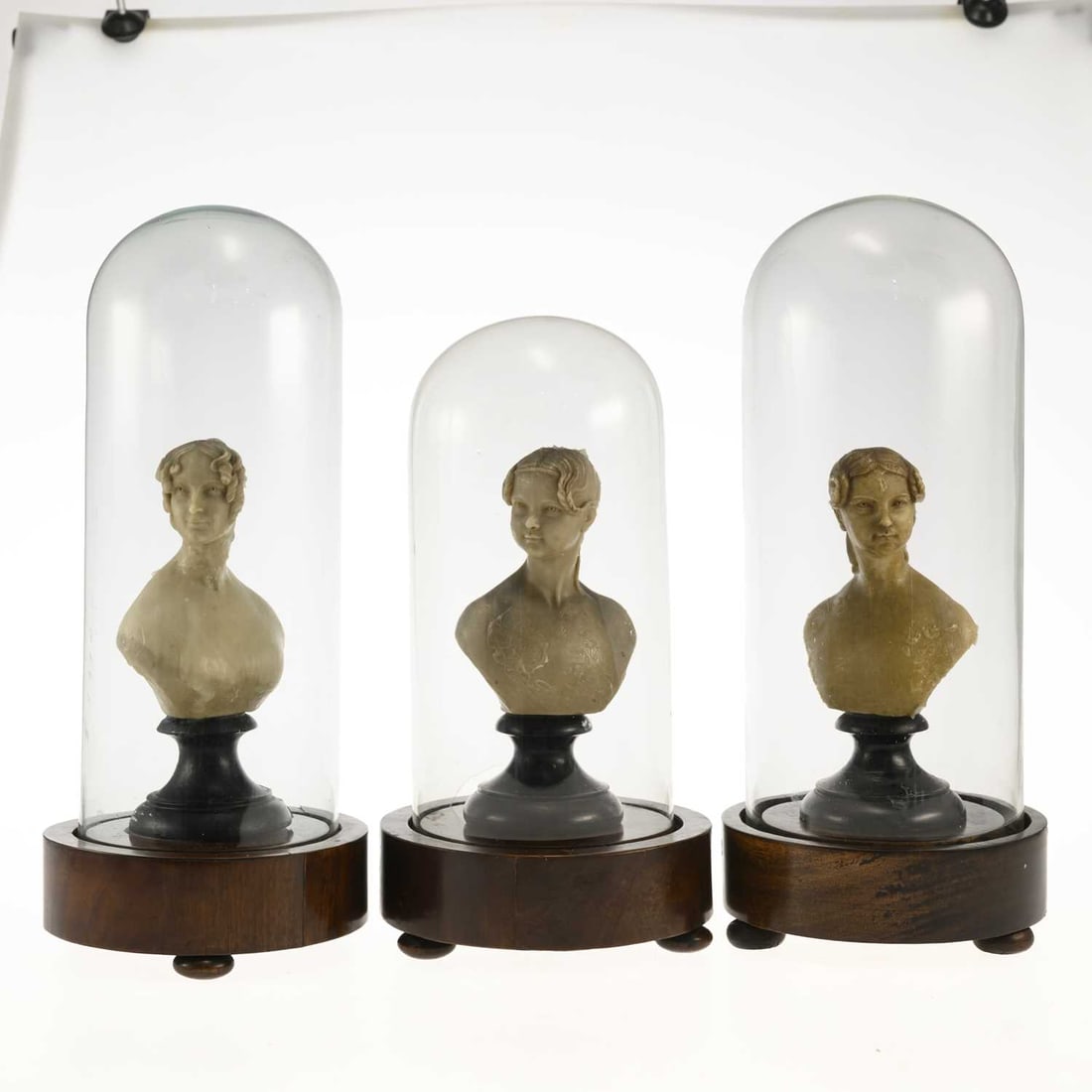 A GROUP OF THREE 19TH CENTURY WAX PORTRAIT BUSTS: A GROUP OF THREE 19TH CENTURY WAX PORTRAIT BUSTS each modelled as a young woman and raised on an ebonised turned socle, beneath a glass dome on a circular mahogany base with bun feet. (3) Tallest 41cm