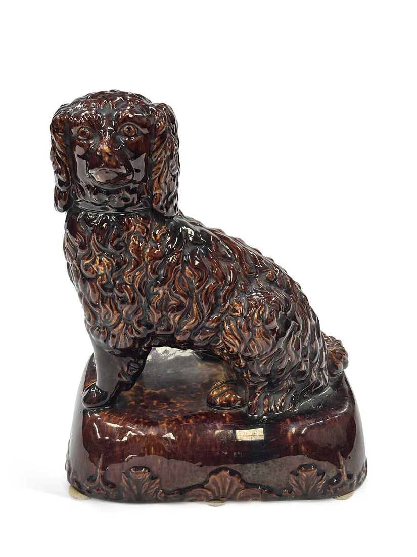 A TREACLE GLAZED POTTERY FIGURE OF A SPANIEL 19TH CENTURY: modelled seated with a combed coat and moulded collar, raised on a shaped rectangular base moulded with shells and scrolls. 29cm high
