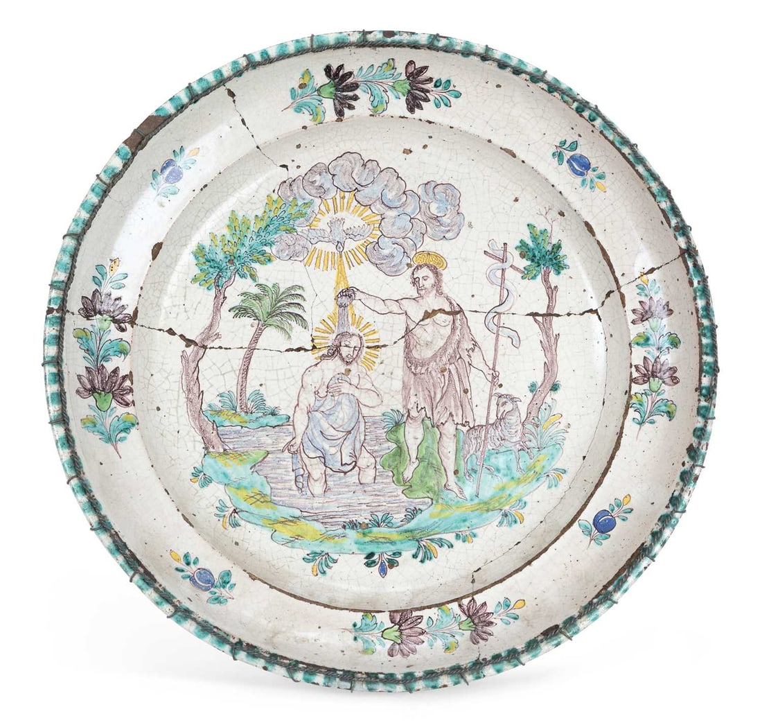 A TIN GLAZED EARTHENWARE CHARGER: painted with the Baptism of Christ, Saint John the Baptist pouring water from a shell as the Holy Spirit descends in a radiance among clouds, within a landscape with palms, the well and border painted