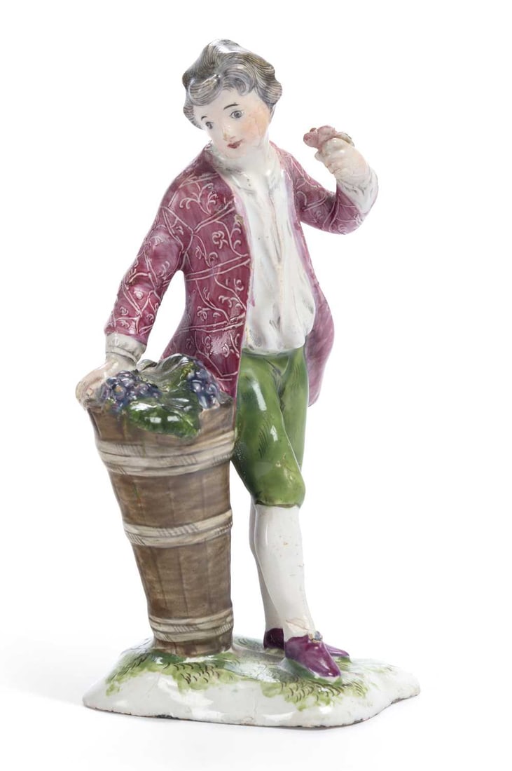 A FRENCH NIDERVILLER FAIENCE FIGURE OF A YOUTH CIRCA 1790: modelled as a young boy in 18th Century attire, with a large basket of grapes, set upon a shaped grassy mound base. 13.3cm high