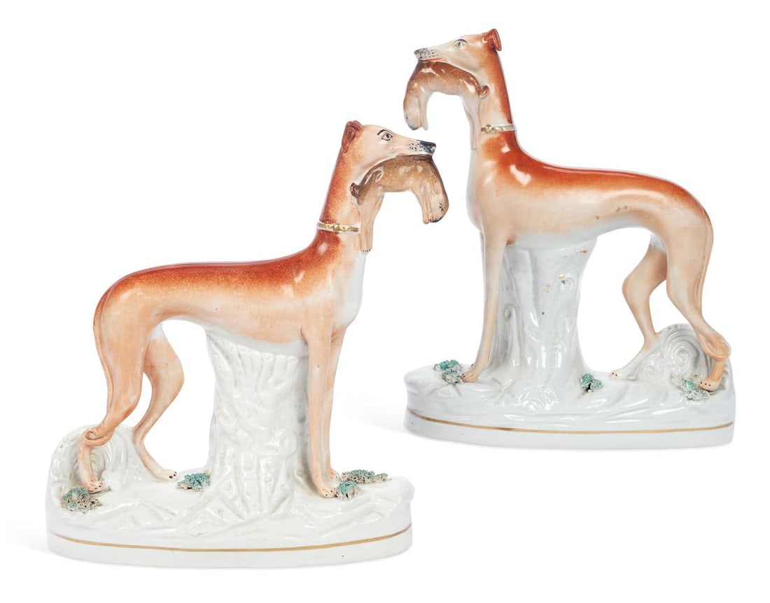 A PAIR OF STAFFORDSHIRE FIGURES OF GREYHOUNDS 19TH CENTURY: each modelled standing before a tree stump with a hare in its mouth, wearing a gilt collar, on an oval base applied with moss and edged in gilt. (2) 28cm high