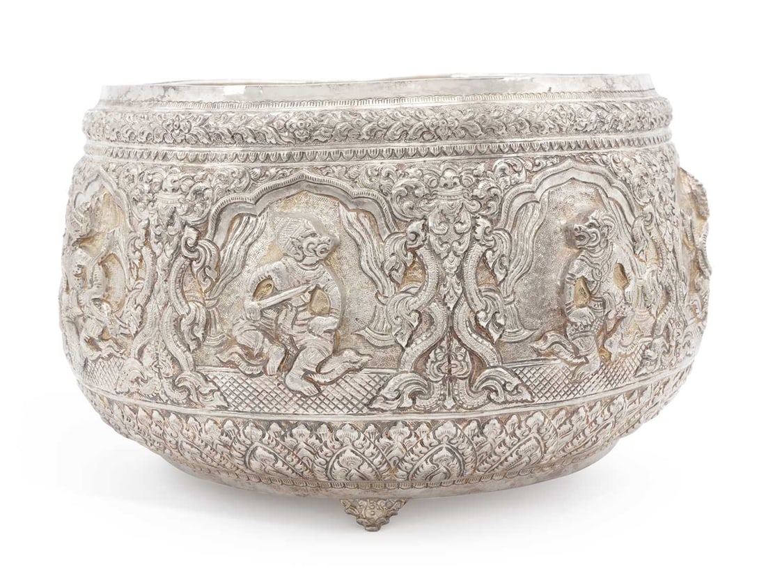 A LARGE BURMESE OR THAI SILVER BOWL EARLY 20TH CENTURY: unmarked, circular with rounded tapering sides, repousse decorated in high relief with seated figures of deities within a decorative leaf and scroll border, raised on three scroll feet. 22.6cm high,