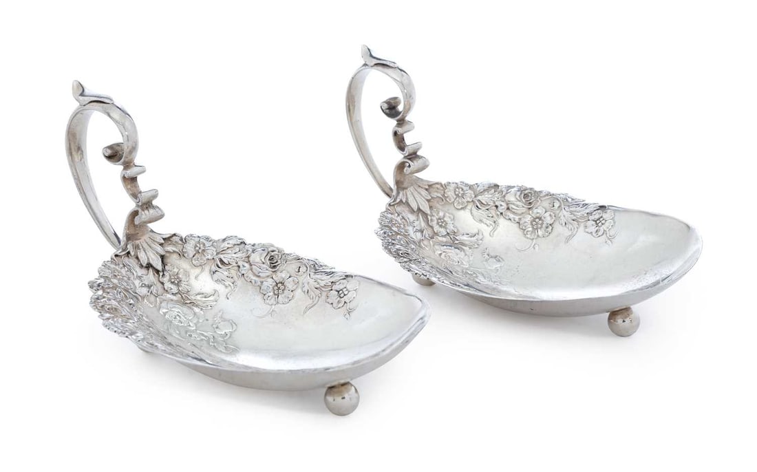 A PAIR OF AMERICAN STERLING SILVER HANDLED DISHES: each of oval form with chased floral motif, scroll shaped handles, raised on three ball feet. (2) 12.5cm long, 6.2cm high, 5.7 troy ounces Good condition overall, signs of surface wear, sticker