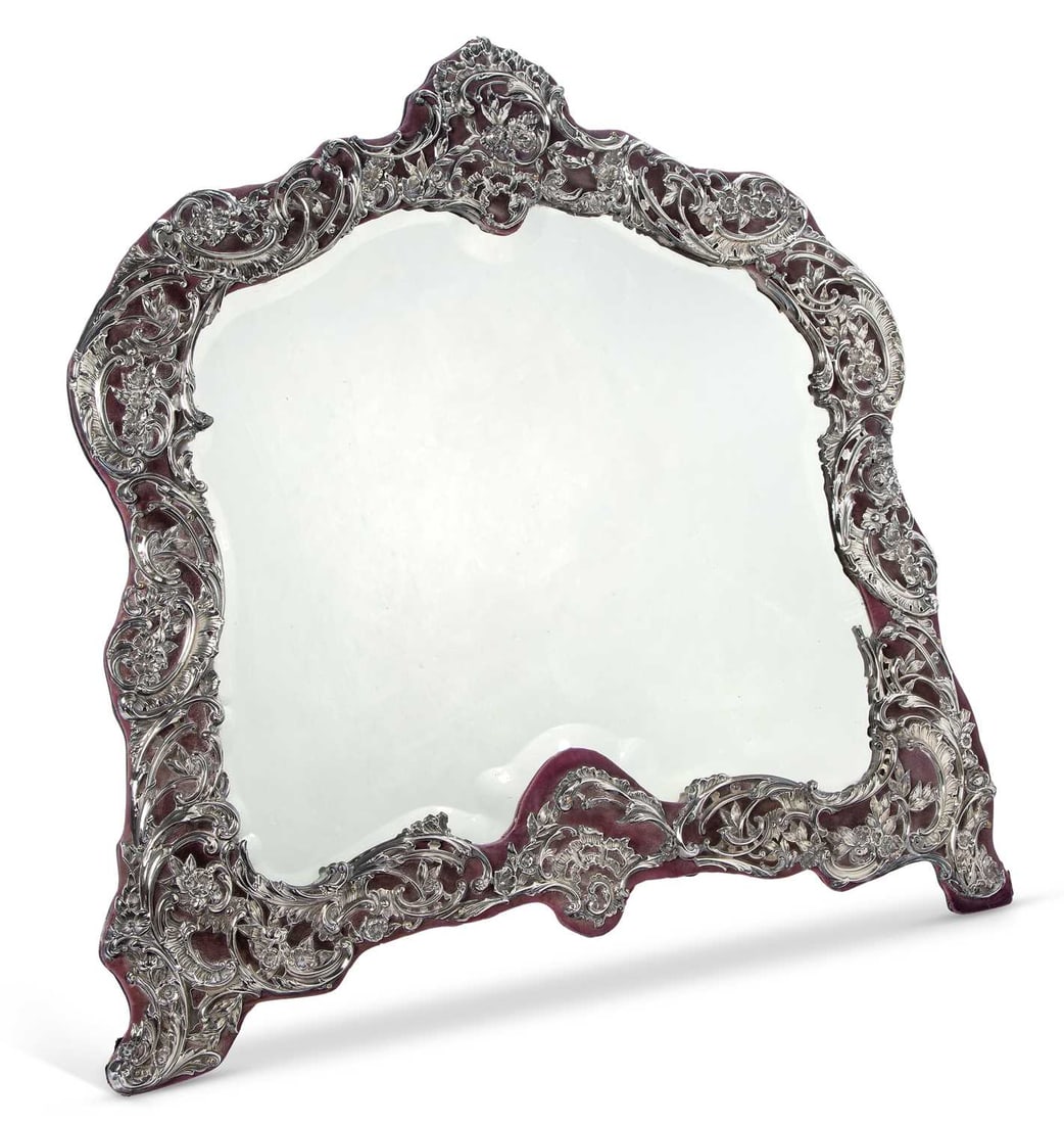 A VERY LARGE EDWARDIAN SILVER-MOUNTED DRESSING TABLE MIRROR: by William Comyns & Sons, London 1906, the shaped arched plate with bevelled edge, within a frame embossed and pierced with rococo scrolls, flowering foliage and cartouches over a velvet ground, to a 