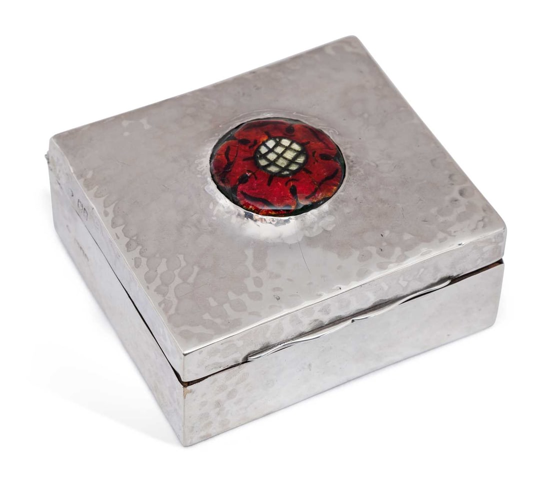 AN EDWARDIAN ARTS AND CRAFTS SILVER AND ENAMEL CIGARETTE BOX: by William Comyns, London 1905, of rectangular form with a hinged cover, with hammered decoration, the shaped lid inset centrally with an enamelled Tudor style red rose, with wooden lining. 8.2cm by 7