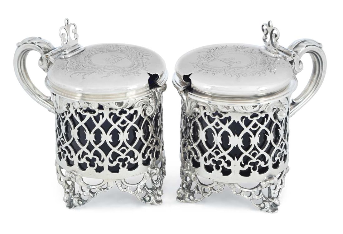 A PAIR OF VICTORIAN SILVER MUSTARD POTS: by Charles Thomas Fox and George Fox, London 1842, of cylindrical form with pierced sides, C-shaped scroll handles and decorative feet, engraved crest within a Rococo cartouche to the hinged cover.