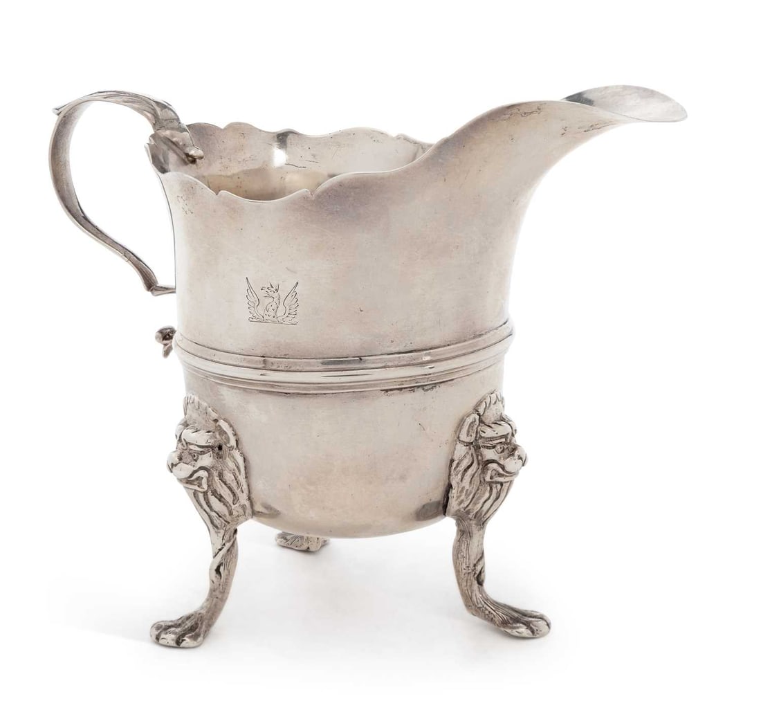 A GEORGE III SILVER CREAM JUG: marks rubbed, probably Irish, of helmet form with shaped rim, reeded girdle, leaf-capped scroll handle, raised on three lion mask and paw feet, engraved with a winged griffin crest. 11.2cm high, 5.4 t
