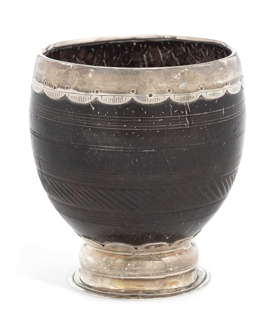 A SILVER-MOUNTED COCONUT CUP LATE 18TH/ EARLY 19TH CENTURY: unmarked, the cup carved with reeded and linear bands, the scallop-edged mounts engraved with linear motif, on a circular spreading foot. 9.5cm high Condition commensurate with age and use, rim