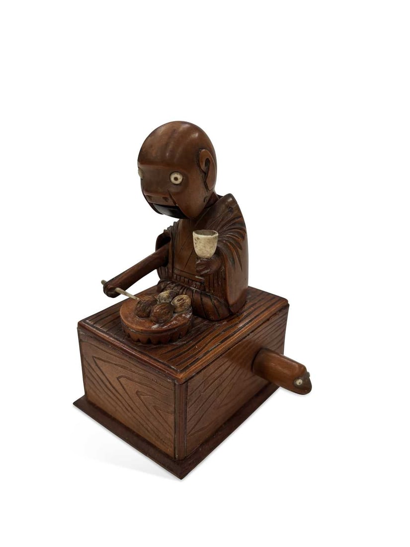A JAPANESE KOBE TOY MEIJI PERIOD: carved as seated figure holding a cup and chopstick, whilst eating from a plate before him, set upon box carved with wood grain, and housing the movement, which is activated by a handle carved as an e