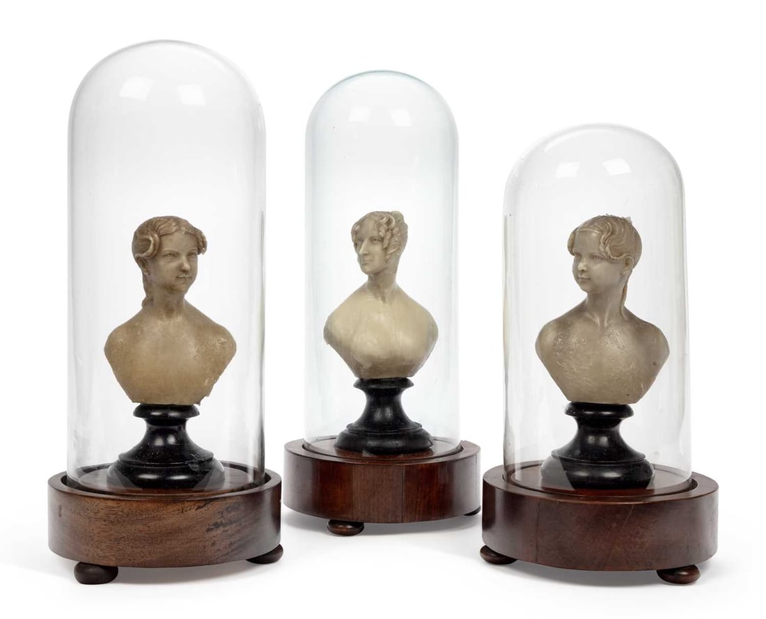 A GROUP OF THREE WAX PORTRAIT BUSTS 19TH CENTURY: each modelled as a young woman and raised on an ebonised turned socle, beneath a glass dome on a circular mahogany base with bun feet. (3) Tallest 41cm high overall