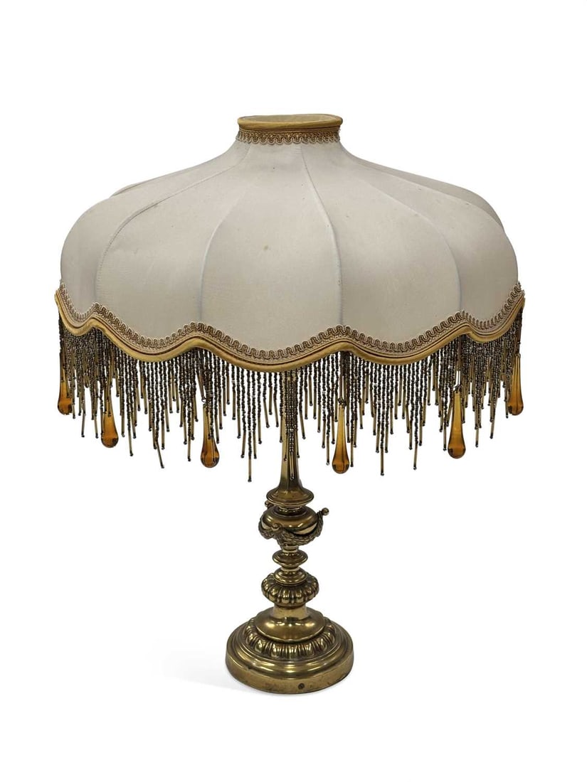 A BRASS TABLE LAMP 20TH CENTURY: the slender tapering column with a knopped and gadrooned stem cast with a ribbon tied swag, on a stepped and gadrooned circular base, with a cream silk shaped shade with an amber and clear glass beade