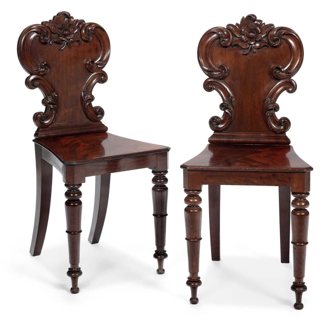 A PAIR OF VICTORIAN MAHOGANY HALL CHAIRS: each with a shaped cartouche back carved with scrolls and centred by a flowerhead, above a solid dished seat, on ring turned tapering front legs and swept rear legs. (2) Height of backs 88cm