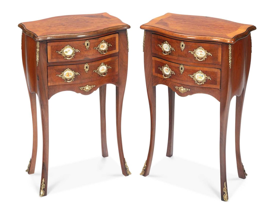 A PAIR OF WALNUT BEDSIDE COMMODES IN THE LOUIS XV TASTE: each with a shaped serpentine top veneered in burr walnut within crossbanding, above two long drawers with gilt metal handles inset with painted porcelain plaques and matching escutcheons, the shaped 