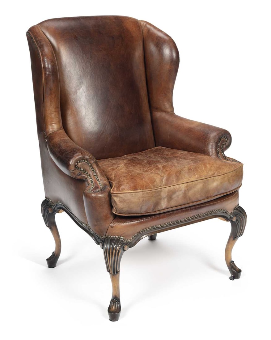 A LEATHER WING ARMCHAIR IN THE GEORGE II TASTE: upholstered in tan leather with close nailing, the arched back between outswept wings above scrolled arms and a loose cushion seat, raised on carved cabriole front legs headed by shells and terminatin