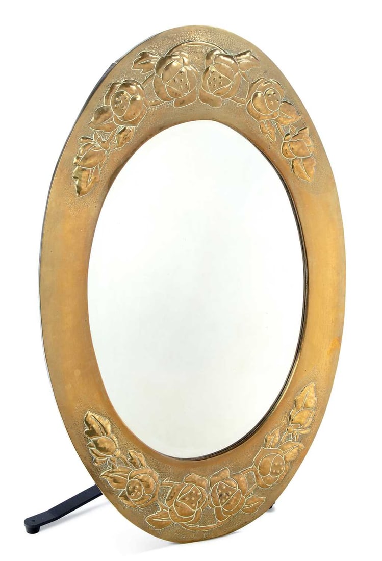 AN ARTS AND CRAFTS BRASS OVAL WALL MIRROR: the frame repousse worked at the top and base with stylised roses and foliage on a punched ground, enclosing a bevelled oval plate. 85cm by 54cm