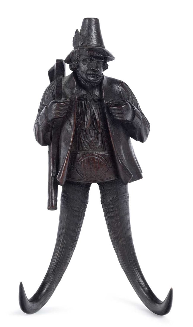 A BLACK FOREST CARVED WOOD WALL HOOK LATE 19TH CENTURY: modelled as a huntsman in a feathered hat and open jacket, a rifle over his shoulder, the legs formed from a pair of chamois horns curving outwards to form the hooks, iron hanging plate to the reverse