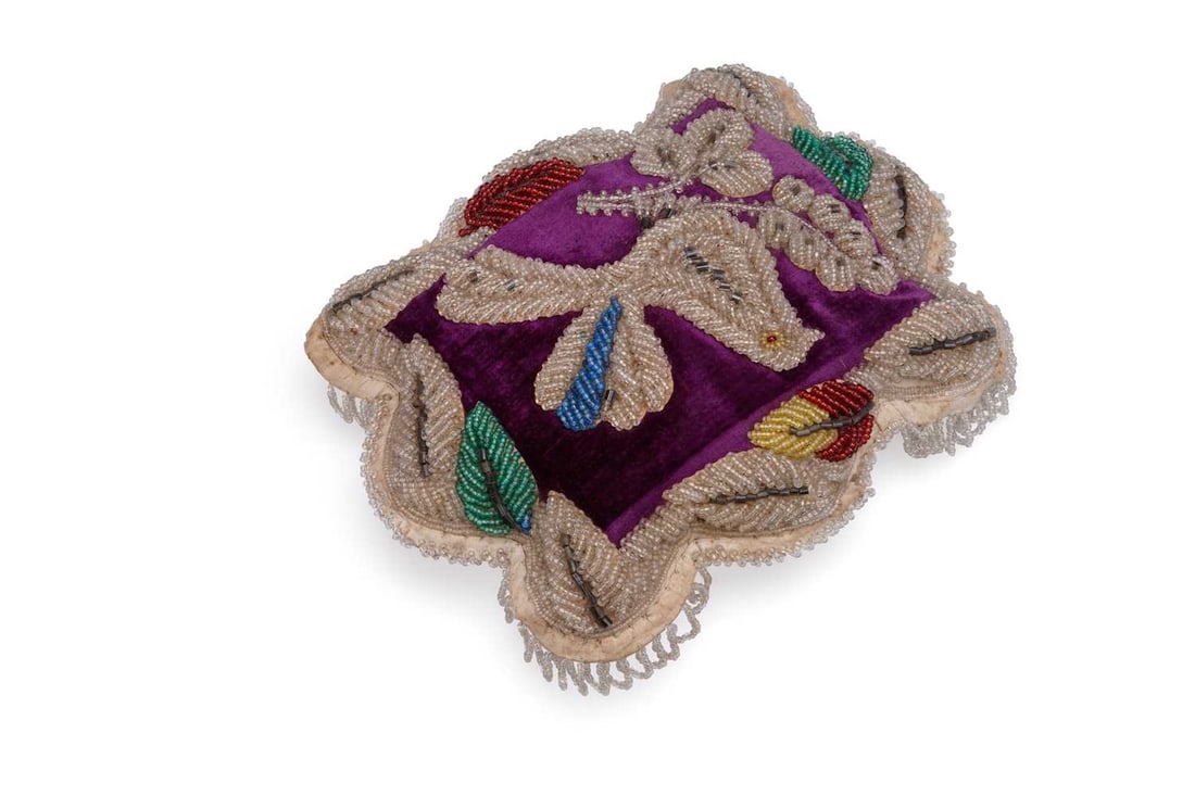 AN IROQUOIS BEADWORK PINCUSHION 19TH CENTURY: AN IROQUOIS BEADWORK PINCUSHION 19TH CENTURY of shaped cushion form, the purple velvet face worked in clear and coloured glass beads with a dove among flowering branches, within a raised petal border