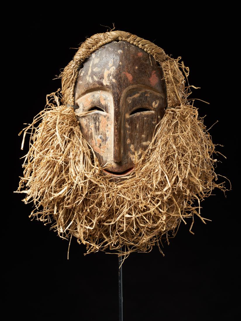 Bira Face Mask from Ituri Region- Tribal Art: A Bira mask from the Ituri Region. This smiling mask is surrounded with straw hair and a straw beard. The mask shows some traces of red and beige pigments. Dimensions approx. A Bira mask from the Itur