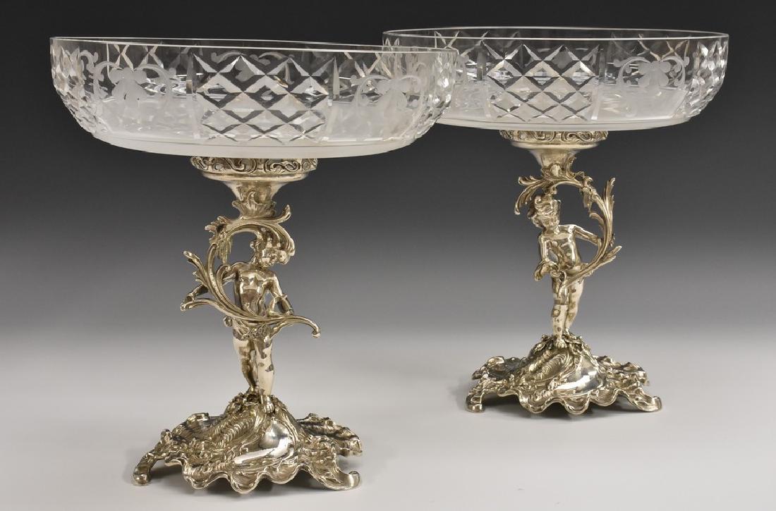 A pair of German silver figural table centre epergnes,: A pair of German silver figural table centre epergnes, each modelled as a scantily clad young boy holding a branch, clear and frosted glass cresting dishes, rocaille bases, 26cm high, 23.5cm diam, hal