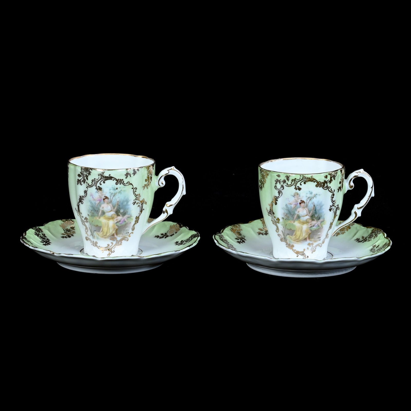 Pair Cups And Saucers, Unmarked Prussia Style: Pair Cups+C125 And Saucers, Unmarked Prussia Style, 2.5 x 4.5 x 4.5 Inches, Green And White With Classic Scene Of Woman And Cherubs With Gold Stencil Highlights, One Saucer Has Rim Chip, 0.4 Pounds.