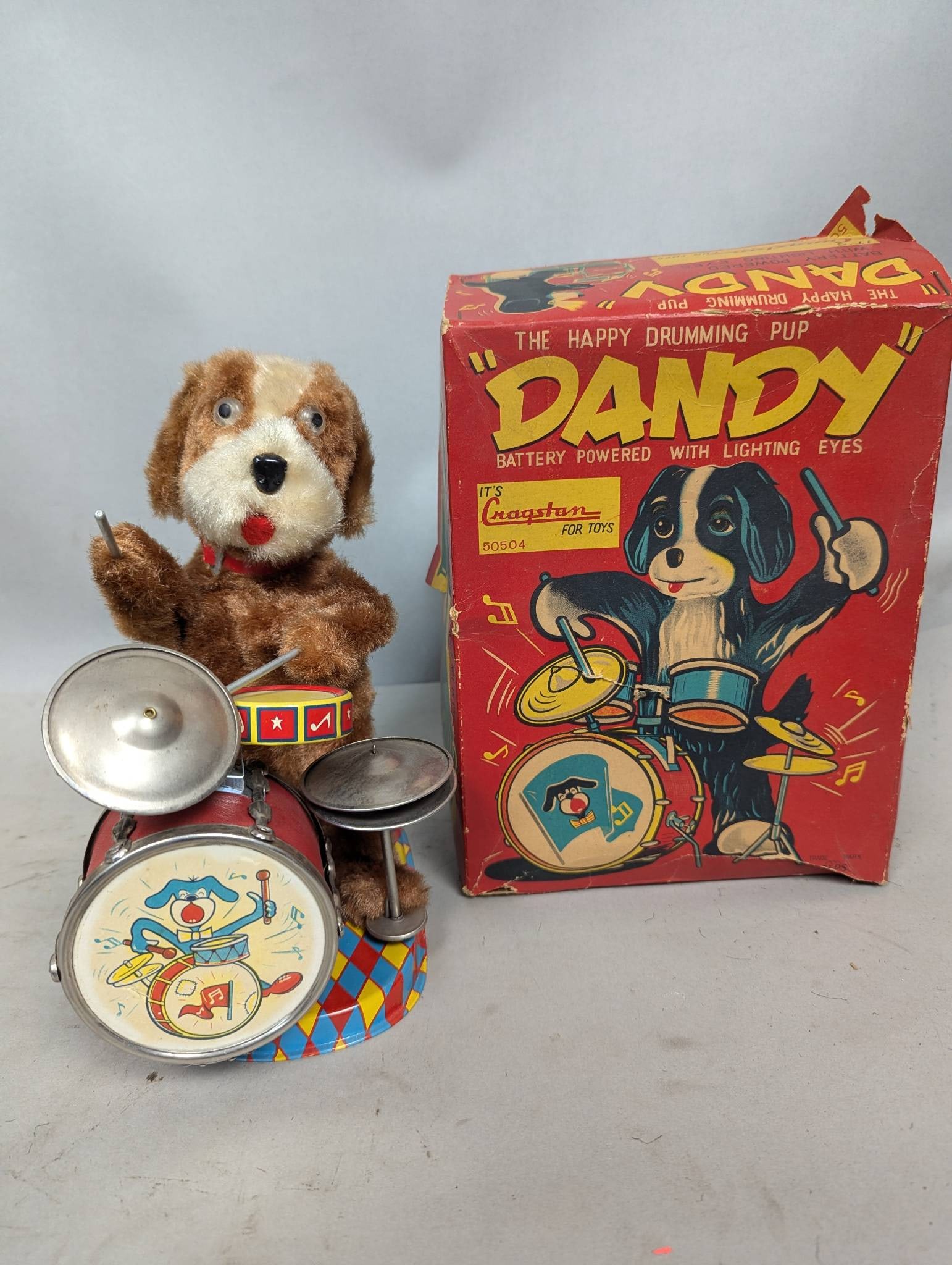 Cragstan Dandy Happy Drumming Pup with Original Box: Battery-powered 'Dandy' Happy Drumming Pup toy manufactured by Cragstan. Constructed with plush material, metal drum set, and colorful carnival-themed base. Includes original box with bright illustrat