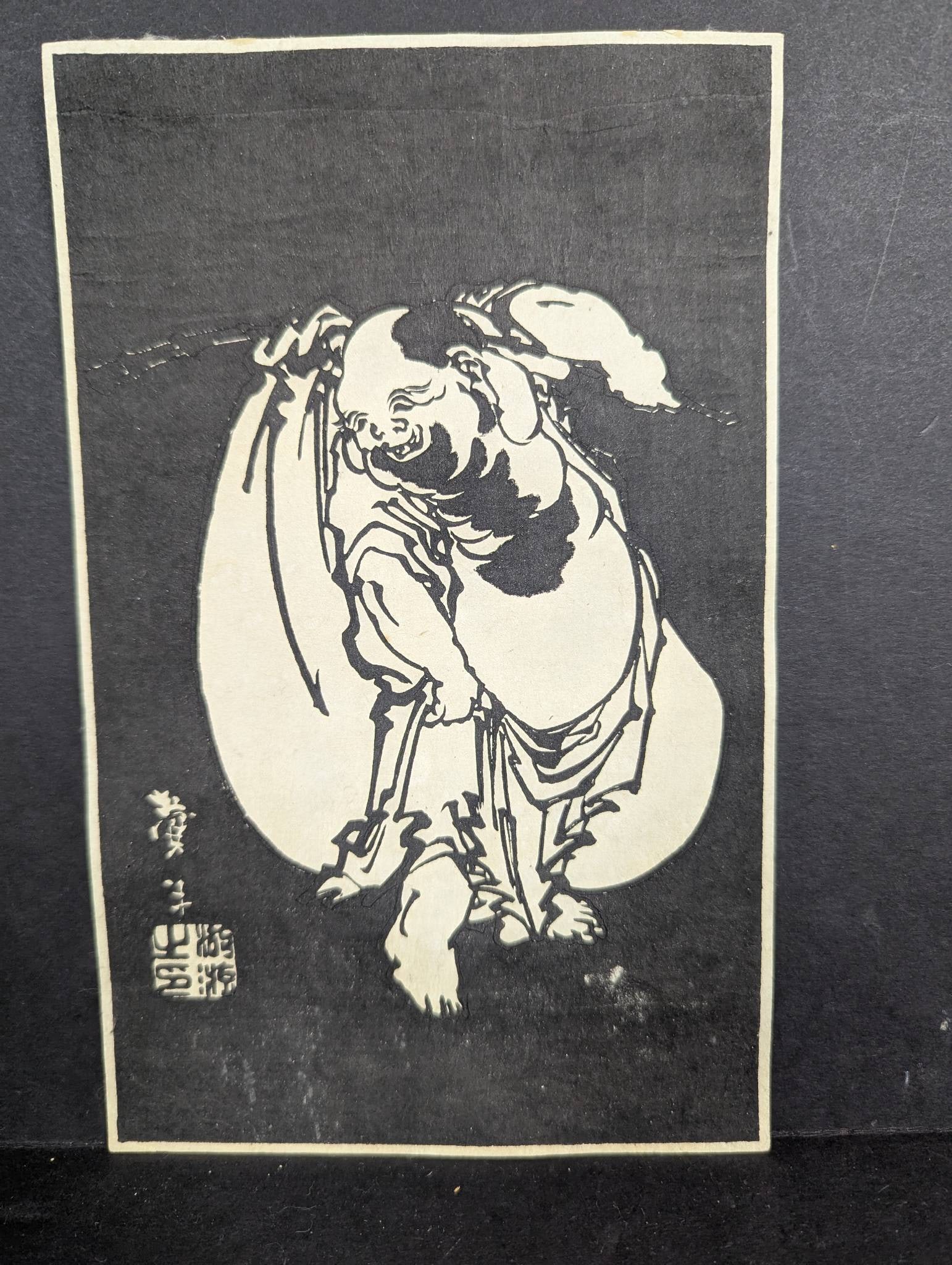 Japanese Woodblock Print by Taito II Depicting Hotei: Japanese woodblock print attributed to Taito II measuring 5 1/2 x 9 inches. Features a depiction of Hotei. The print has a repaired top tear and shows wear. There is tape residue on the reverse side.