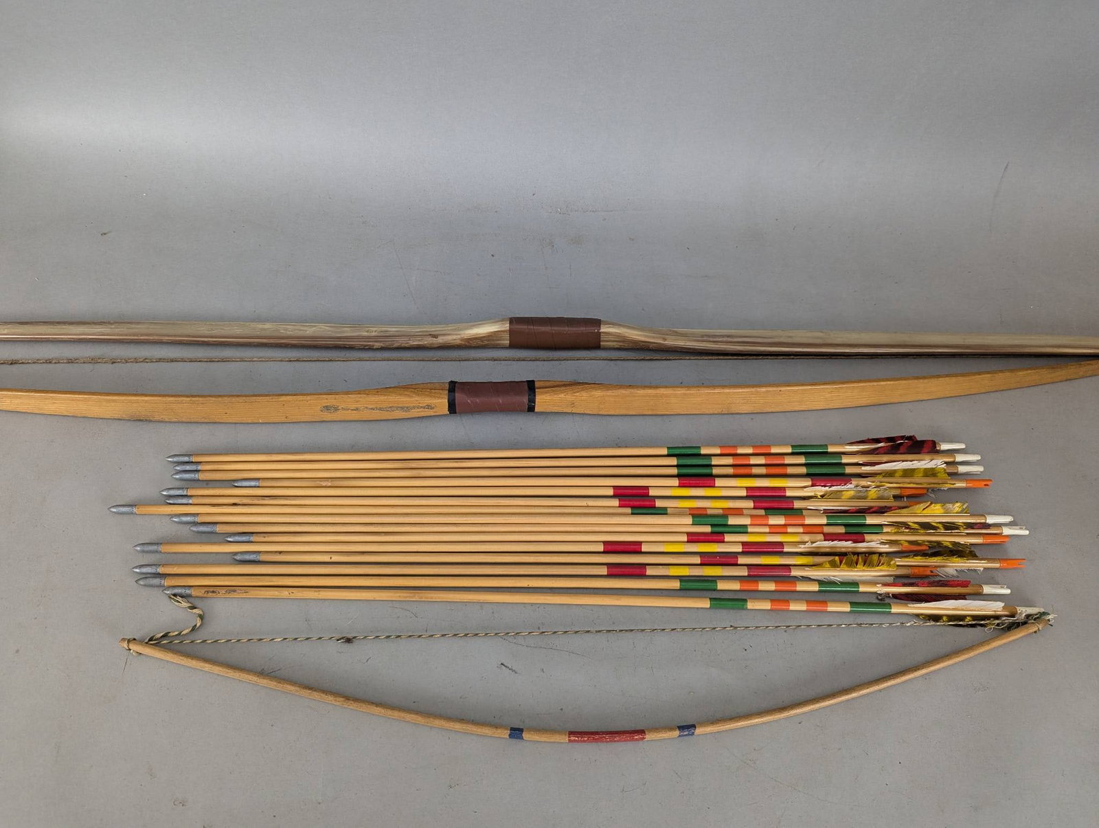 Set of Vintage Wooden Longbows and Arrows with Decorative Fletching: Set of three longbows measuring approximately 30 inches, 54 inches, and 60 inches in length, accompanied by a collection of wooden arrows with decorative feathered fletching in various vibrant colors.