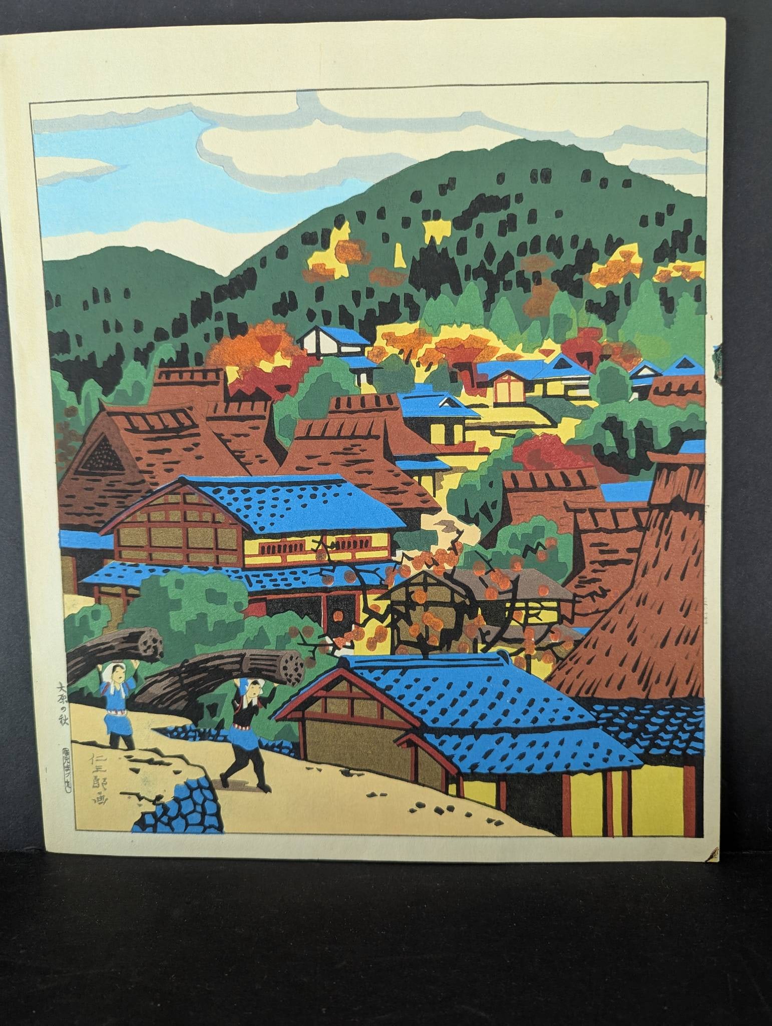 Japanese Woodblock Print by Ito Nisaburo Depicting Autumn in Ohara: Colorful Japanese woodblock print by Ito Nisaburo (1905-2001), titled Autumn in Ohara. The scene depicts a charming village with traditional thatched-roof houses amidst autumn foliage, set against a h