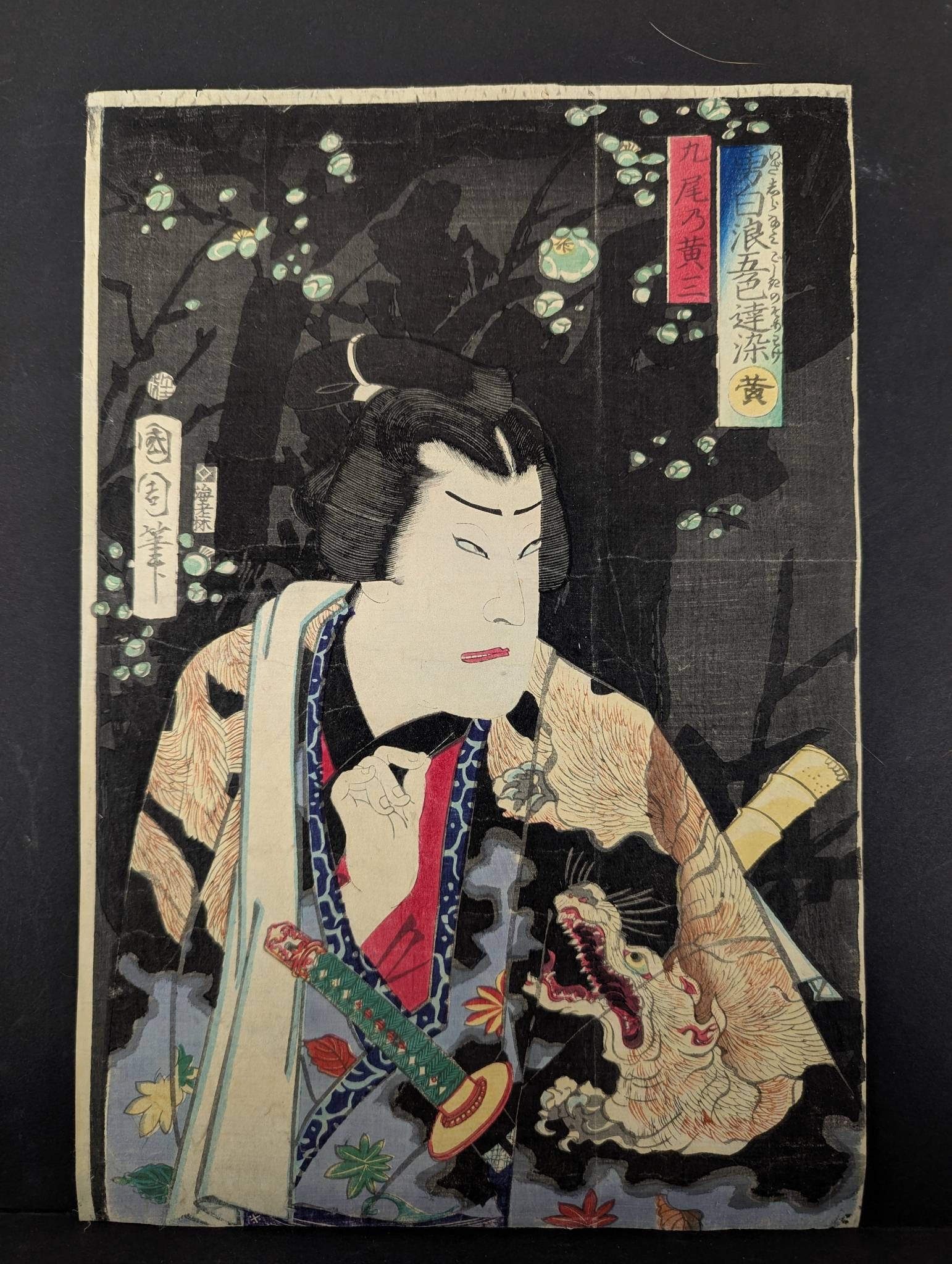 Japanese Woodblock Print by Kunichika Featuring Samurai Scene: Original Japanese woodblock print by Toyohara Kunichika (1835-1900), portraying a samurai dressed in a beautifully patterned kimono holding a katana. The design is set against a dark background adorne
