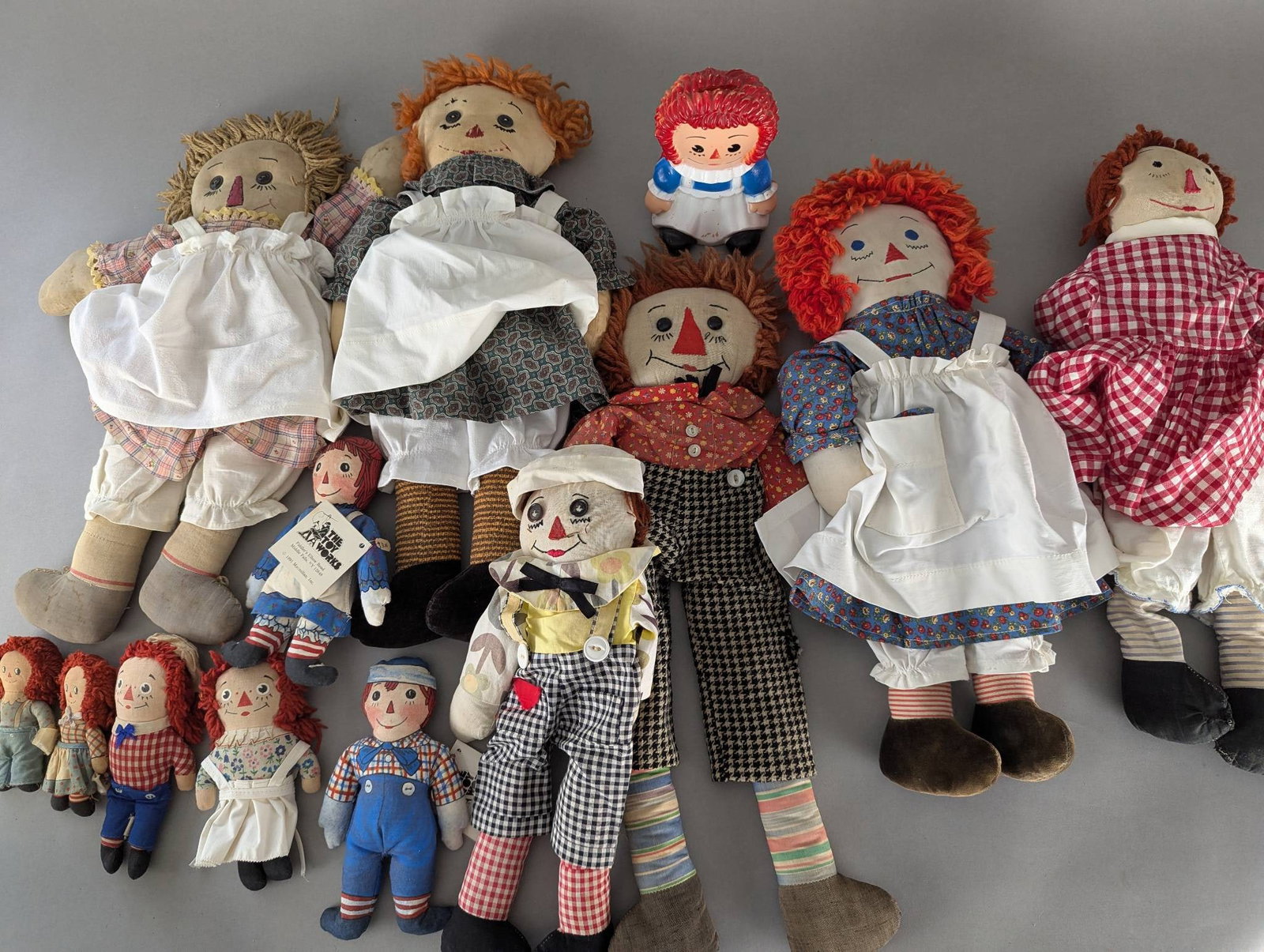 Collection of Raggedy Ann and Andy Dolls in Various Sizes and Styles: A collection of Raggedy Ann and Andy dolls featuring various sizes, materials, and styles. Included are several cloth dolls with traditional red yarn hair and embroidered details, some of which are dr
