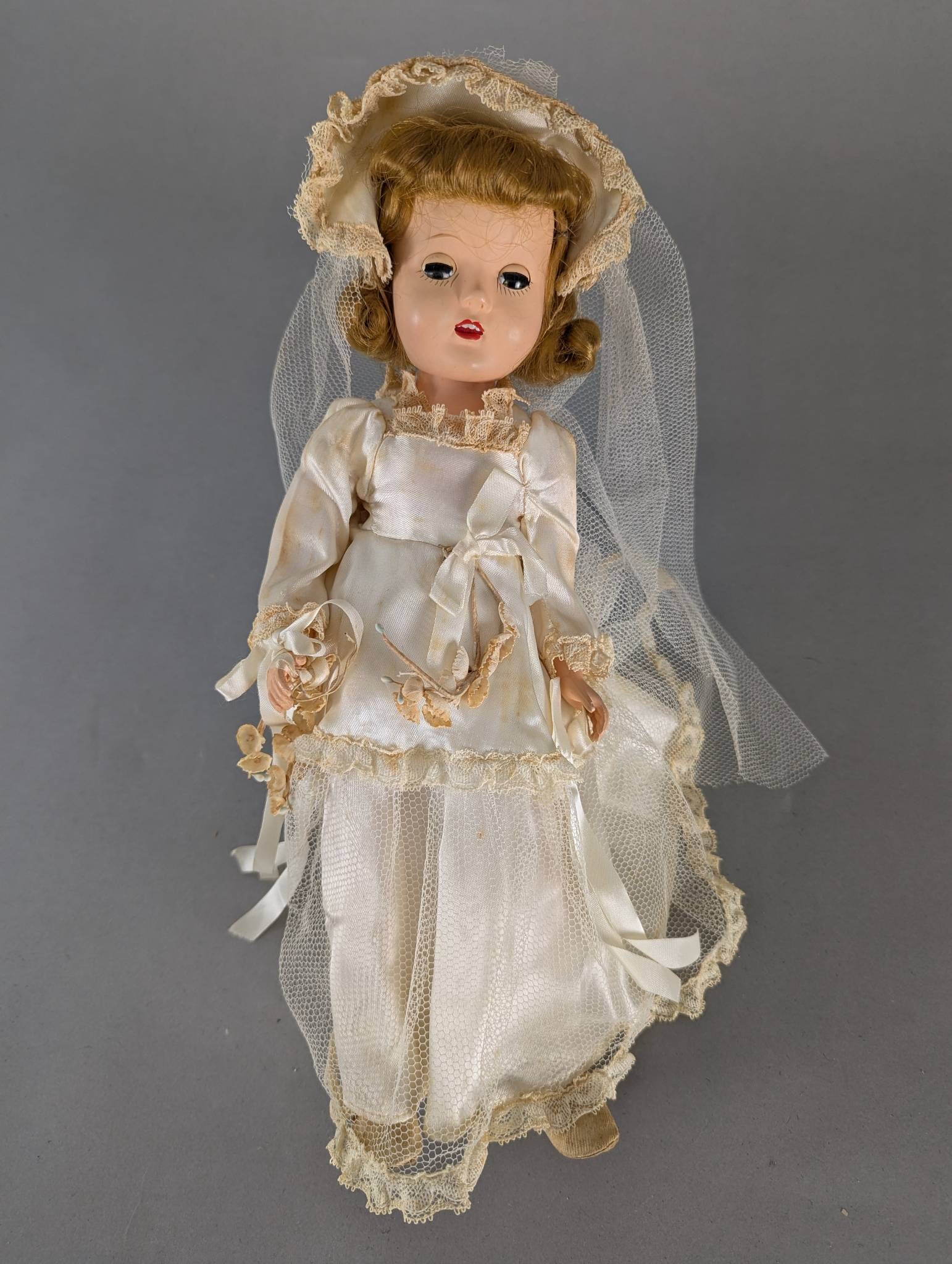 Effanbee Bride Doll in Wedding Dress Made in USA: Effanbee bride doll featuring a detailed white satin wedding dress with lace and tulle detailing. The doll has a painted face with stylistic features, curled hair, and is dressed in an intricately des
