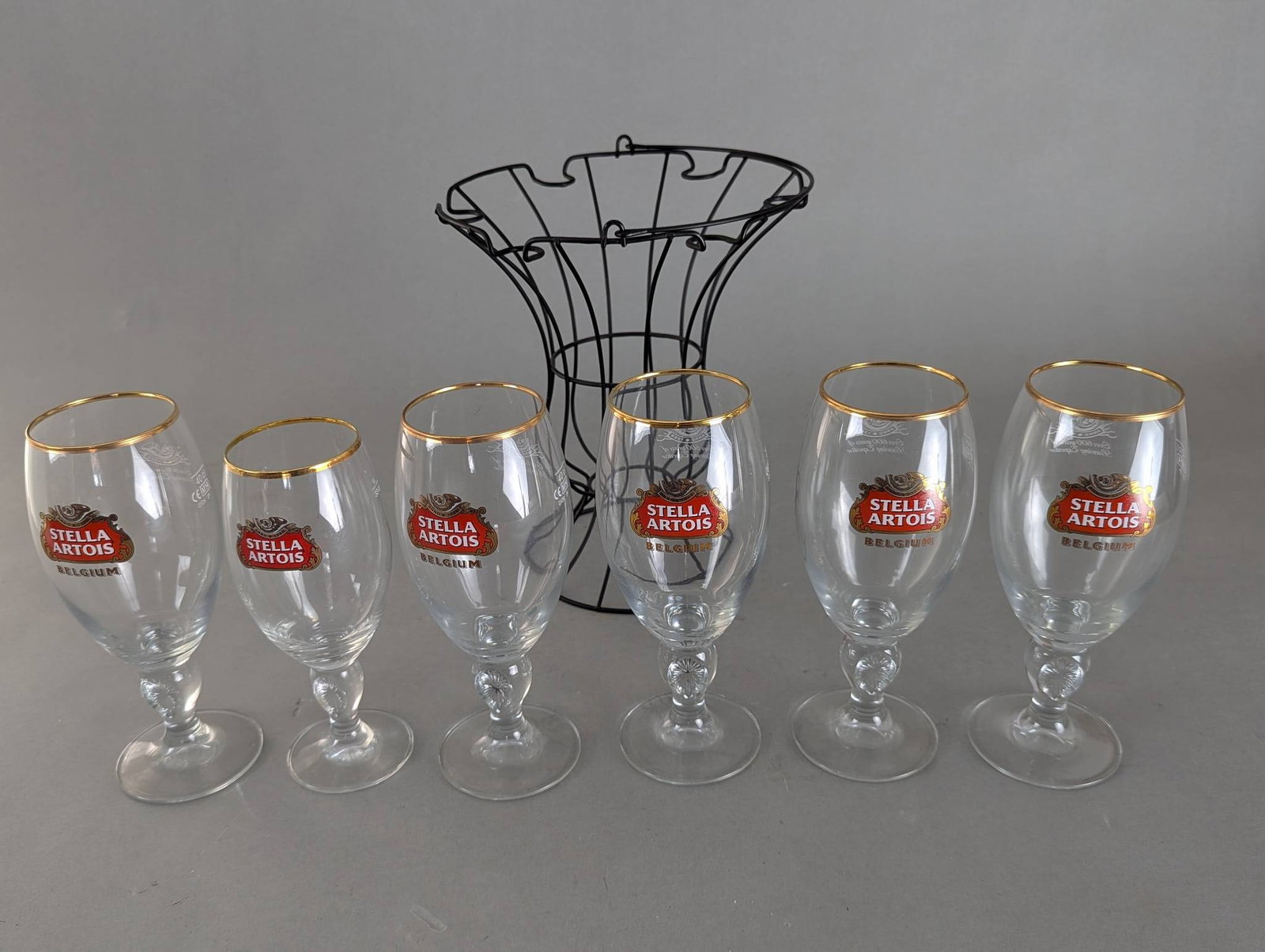 Set of 6 Stella Artois Glasses with Gold Rims and Wire Rack: Set of six Stella Artois branded glasses featuring classy gold rims above their beautifully designed bowls. Each glass is adorned with the official Stella Artois logo and the word Belgium, showcasing 
