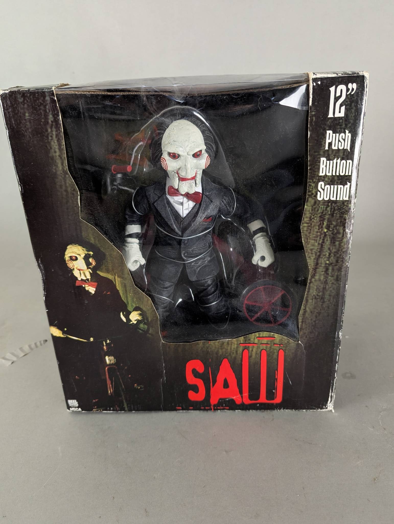 NECA 12 Inch Saw Figure with Push Button Sound Feature New: NECA collectible figure from the Saw franchise. Stands 12 inches tall and features a push-button sound mechanism. Still in the original packaging, which measures approximately 12 high x 13 wide x 8 de