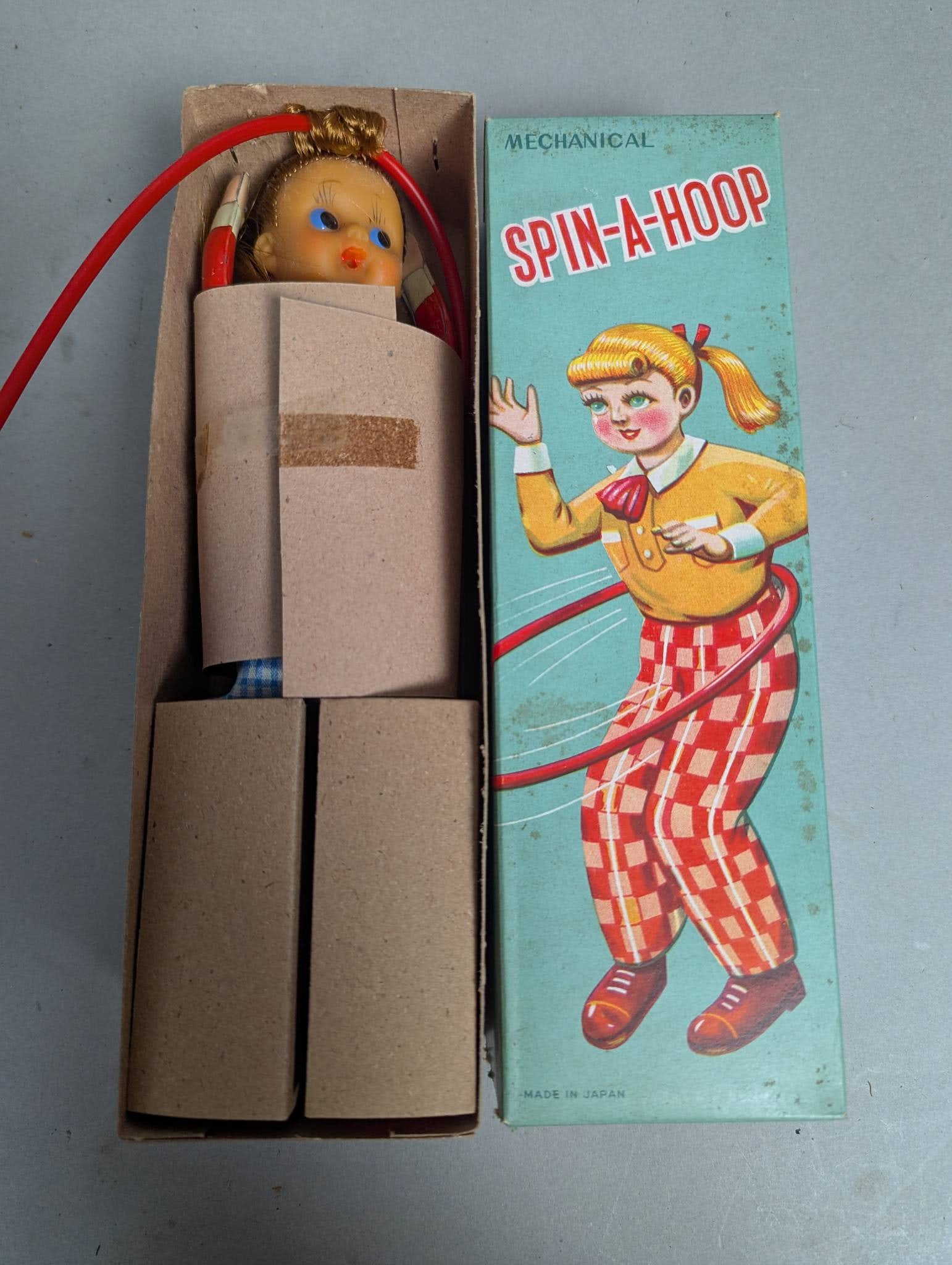 Mechanical Spin A Hoop Toy Made in Japan from 1950s: 1950s mechanical Spin A Hoop toy made in Japan. The toy includes a figure dressed in colorful attire holding a red hula hoop and comes in its original illustrated cardboard box measuring approximately