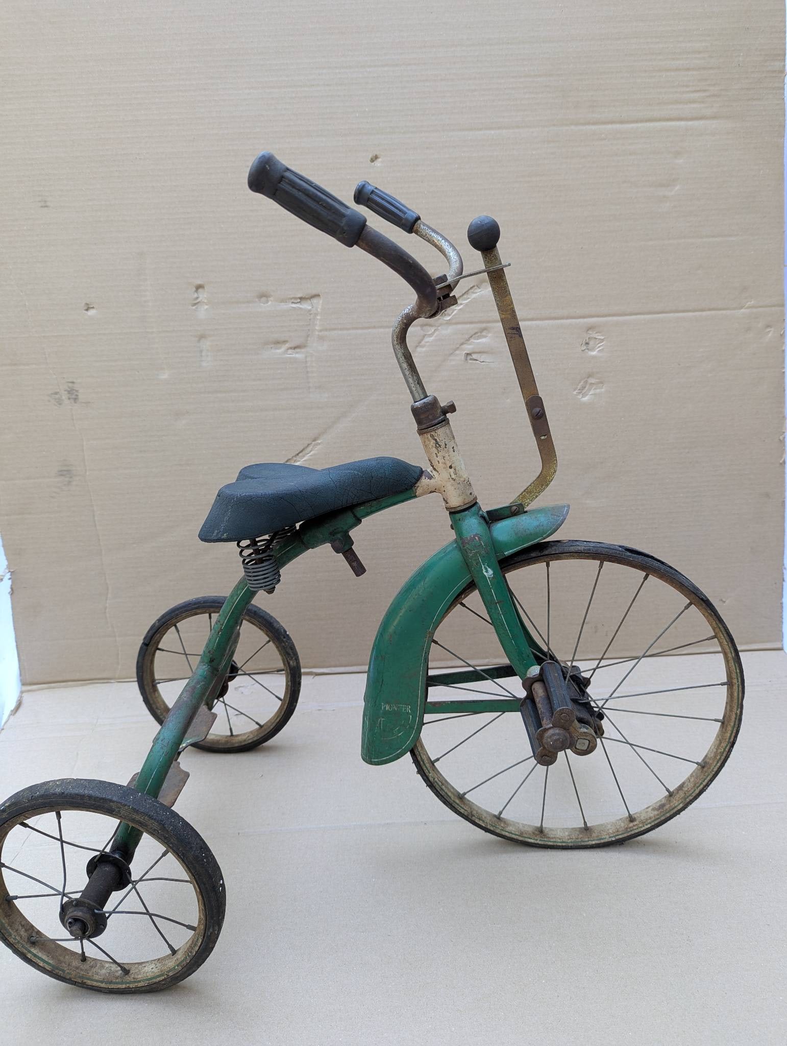 Pioneer Tricycle from the 1930s with Rare Hand Brake: Pioneer tricycle from the 1930s featuring a rare hand brake mechanism. The frame is painted green, and the design includes a curved handlebar with rubber grips, a cushioned saddle, and a solid metal f