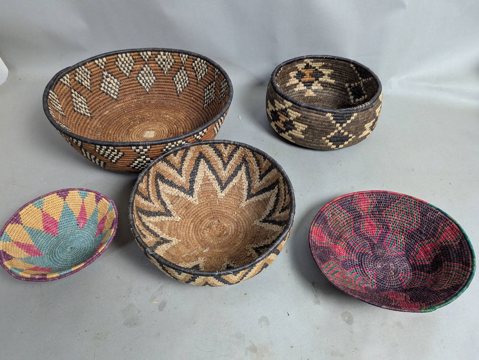 Five Woven Decorative Baskets with Geometric Patterns: Five handwoven decorative baskets, each featuring unique geometric patterns. The largest basket measures 5 high by 15 in diameter. The baskets exhibit a combination of earthy tones and vibrant colors,