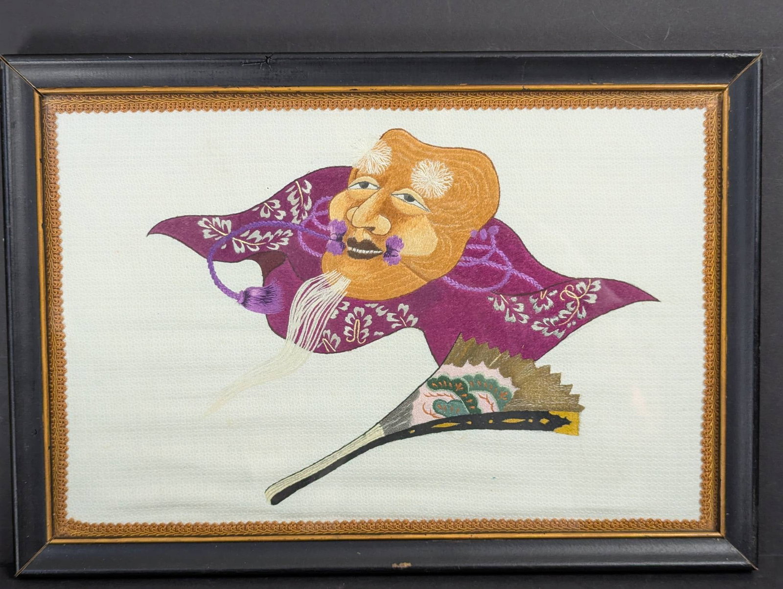 Framed Japanese Noh Mask Embroidery Depicting Okina Mask and Fan: Framed embroidery featuring a Japanese Noh theater Okina mask and a traditional folding fan. The Okina mask symbolizes prosperity and longevity, being one of the central characters in Noh theater. The