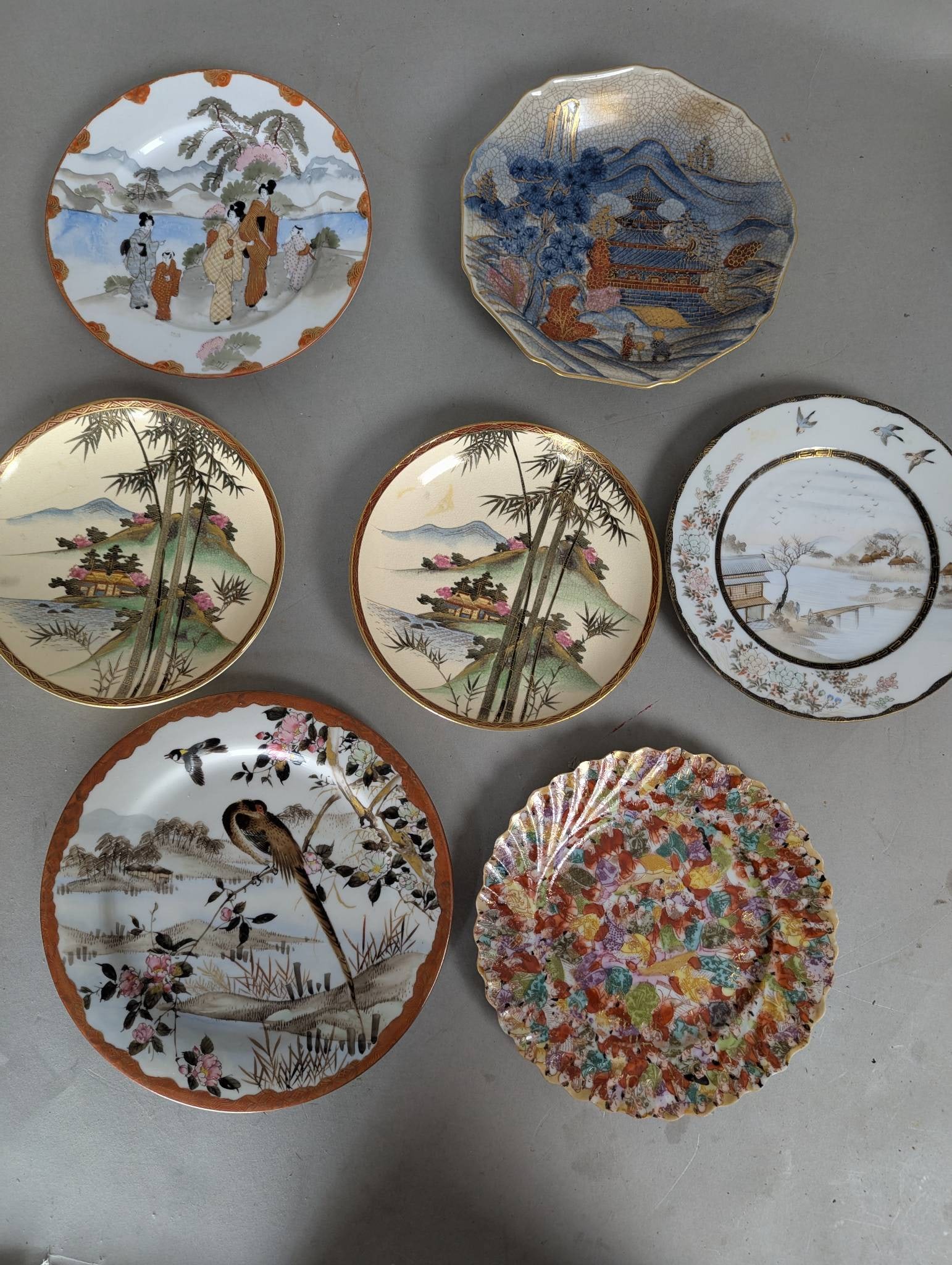 Seven Japanese Porcelain Plates with Hand-Painted Scenes: Seven Japanese porcelain plates featuring various hand-painted designs inspired by traditional Japanese art and culture. The plates depict scenes such as landscapes, bamboo forests, birds, floral patt