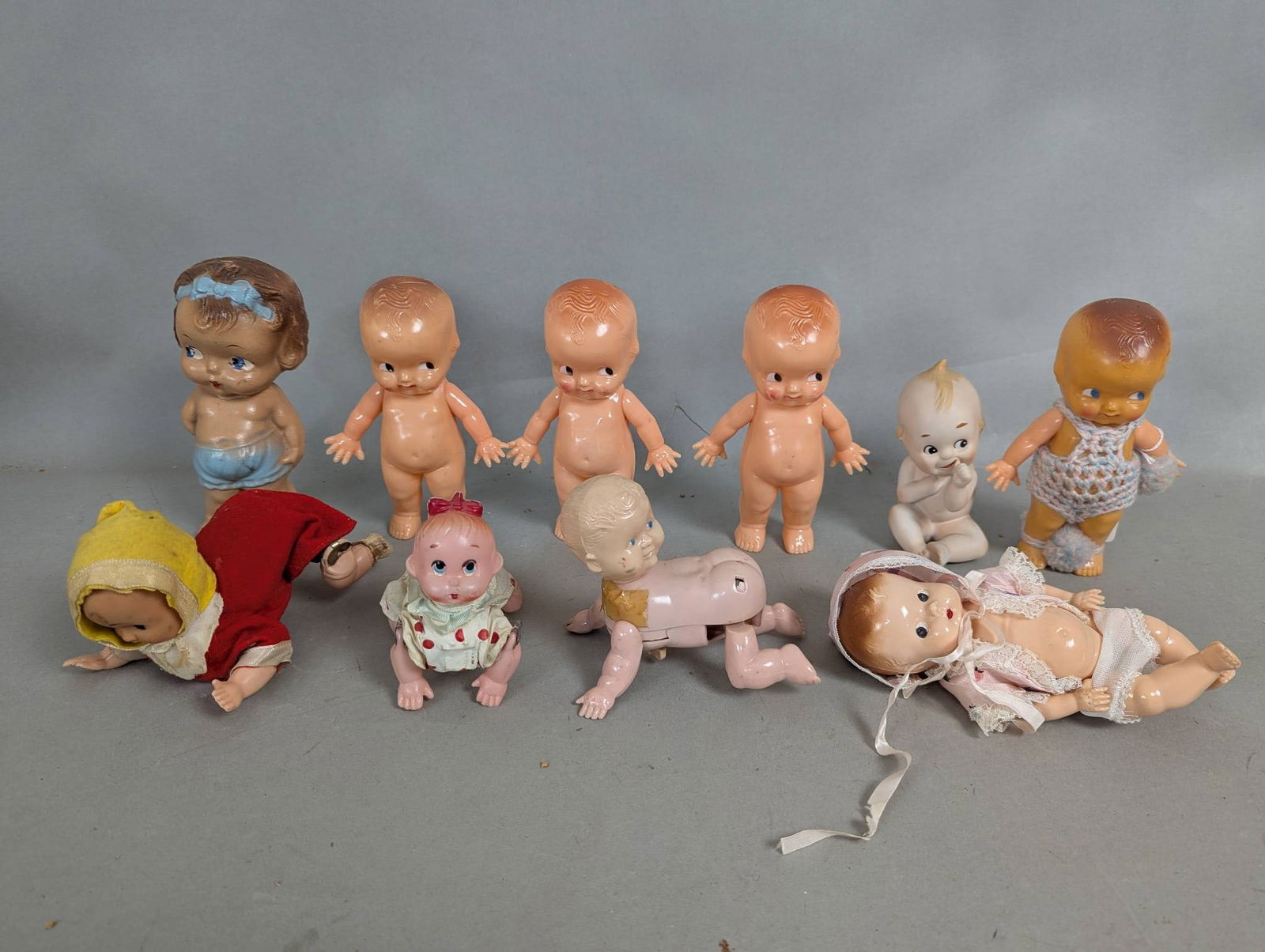 10 Vintage Baby Dolls Made of Hard Plastic with Wind Up Mechanisms: 10 vintage baby dolls crafted from hard plastic. The collection includes a variety of standing and crawling dolls, some of which are wind-up toys. Most dolls have painted features and various outfits,