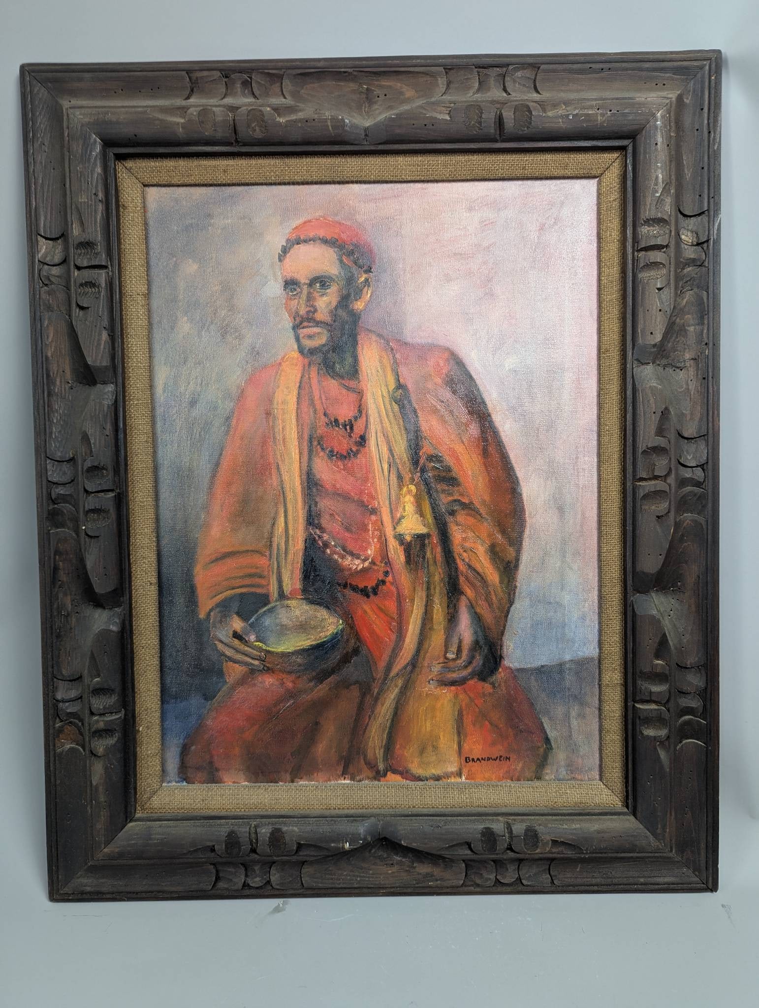 Painting of Indian Sadhu Signed Brandwein in Ornate Wooden Frame: Oil painting depicting an Indian sadhu, created and signed by the artist Brandwein. The artwork portrays a seated figure dressed in traditional orange-toned attire adorned with necklaces, holding a bo