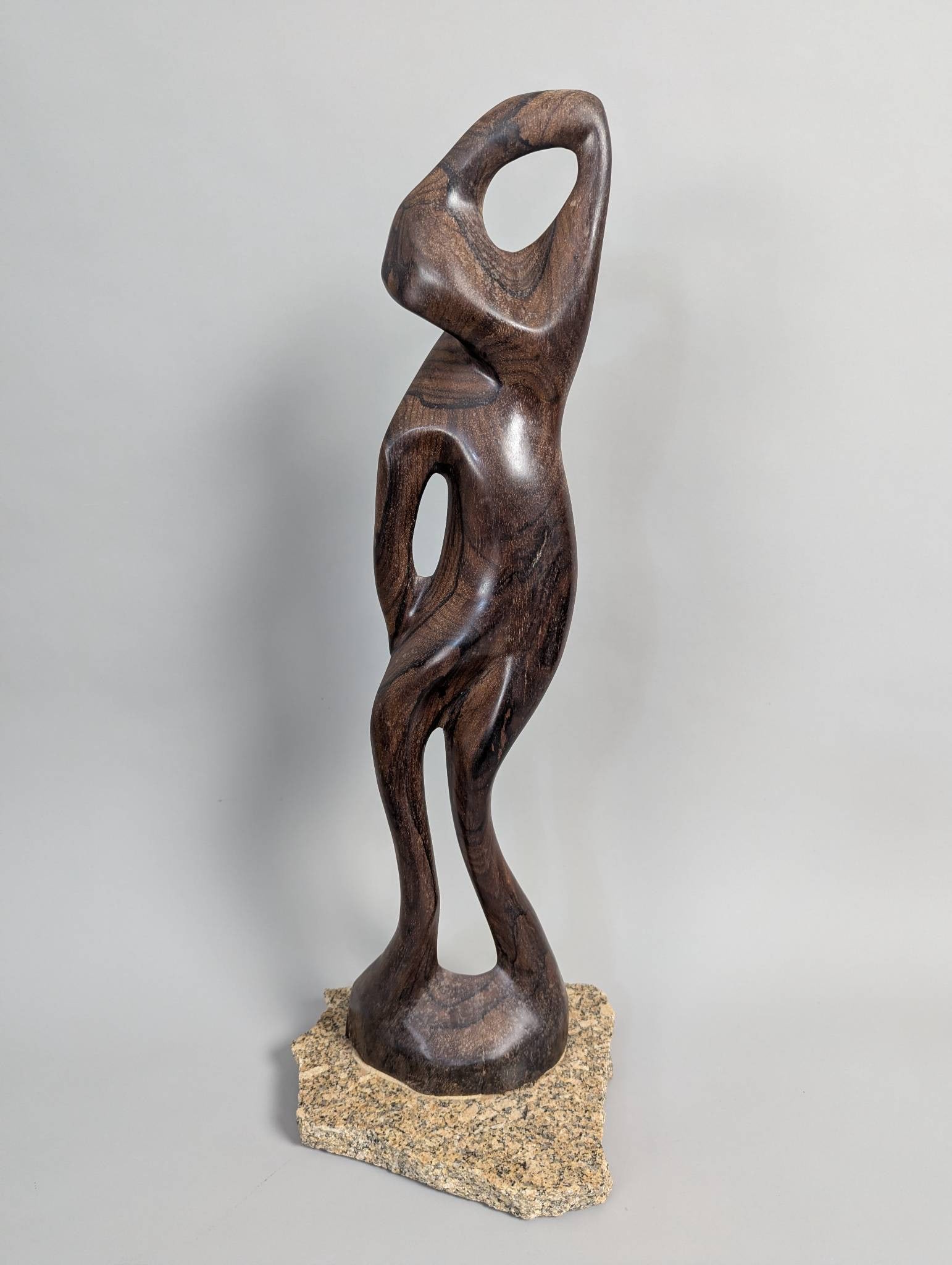 Makonde Hand-Carved Abstract Wood Sculpture on Granite Base: A hand-carved sculpture from a single piece of dark, rich wood, styled in an abstract and flowing design characteristic of the Makonde people of Tanzania. This piece features open spaces within its co