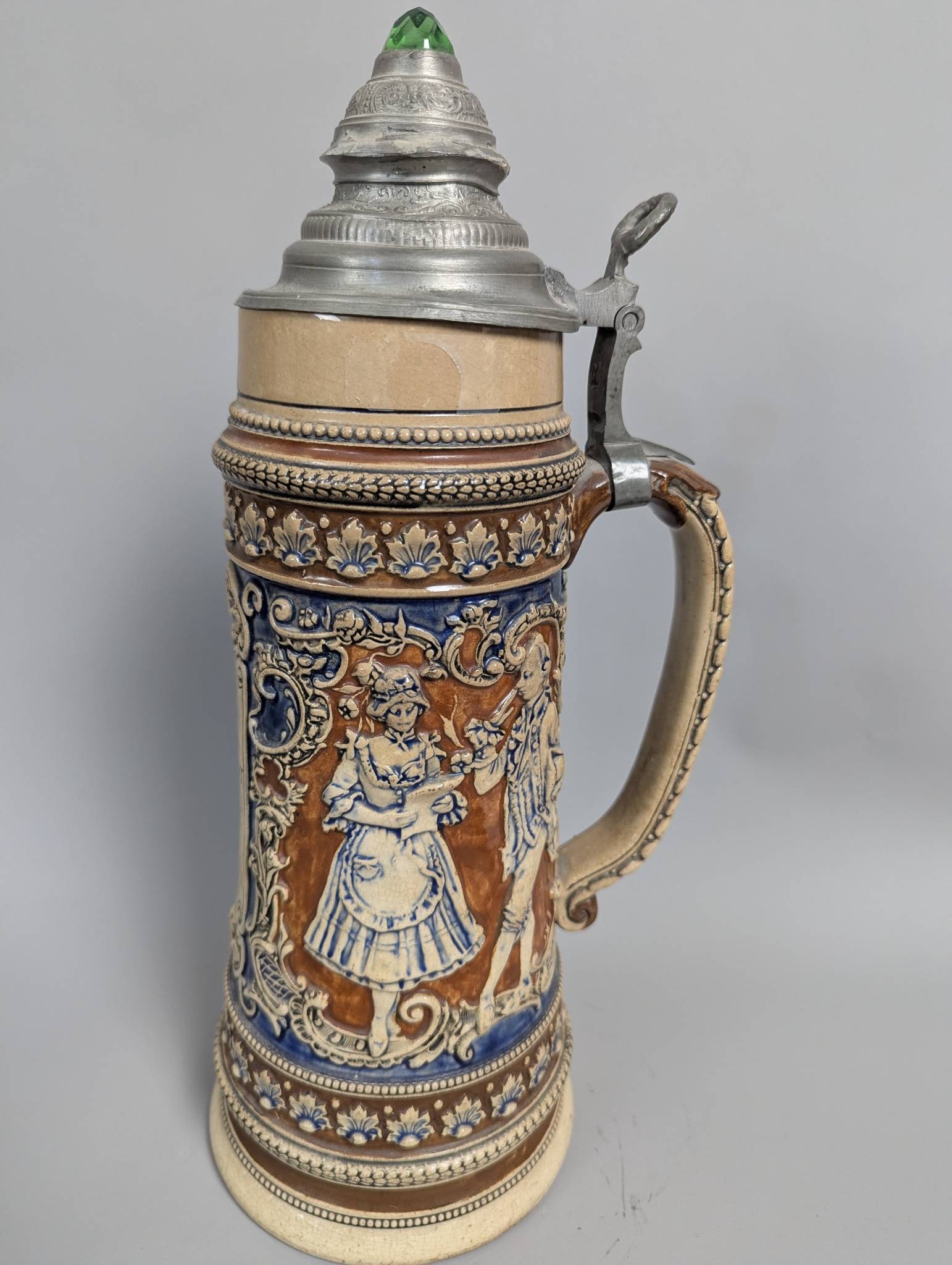 German Stoneware Beer Stein with Pewter Lid and Raised Relief Design 2 Liter: A German beer stein crafted from ceramic stoneware, standing 15 inches high with a 5-inch diameter and a 2-liter capacity. The stein features a pewter lid with a green glass embellishment at the top.