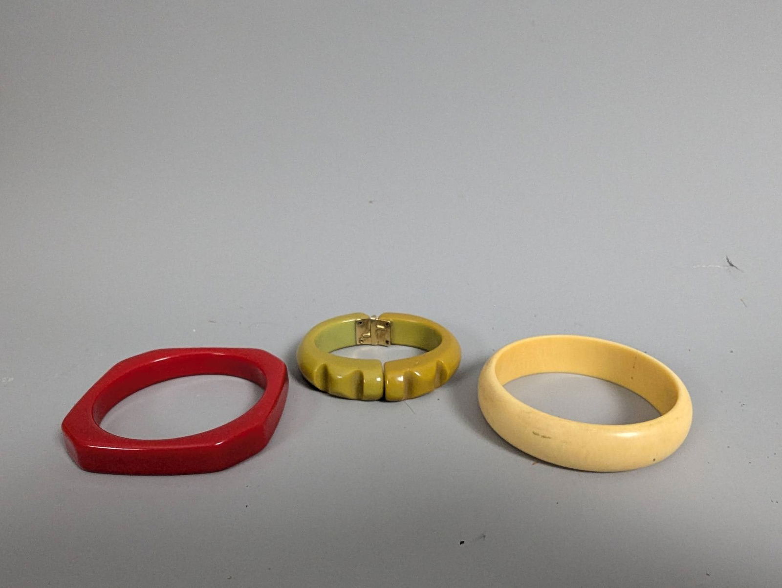 Vintage Bakelite and Lucite Bracelets Including Geometric and Hinged Designs: Three vintage bracelets, potentially made of Bakelite and Lucite. The set includes: a red bracelet with a geometric octagonal shape; a yellow-green carved hinged "clamper" bracelet featuring a brass-t
