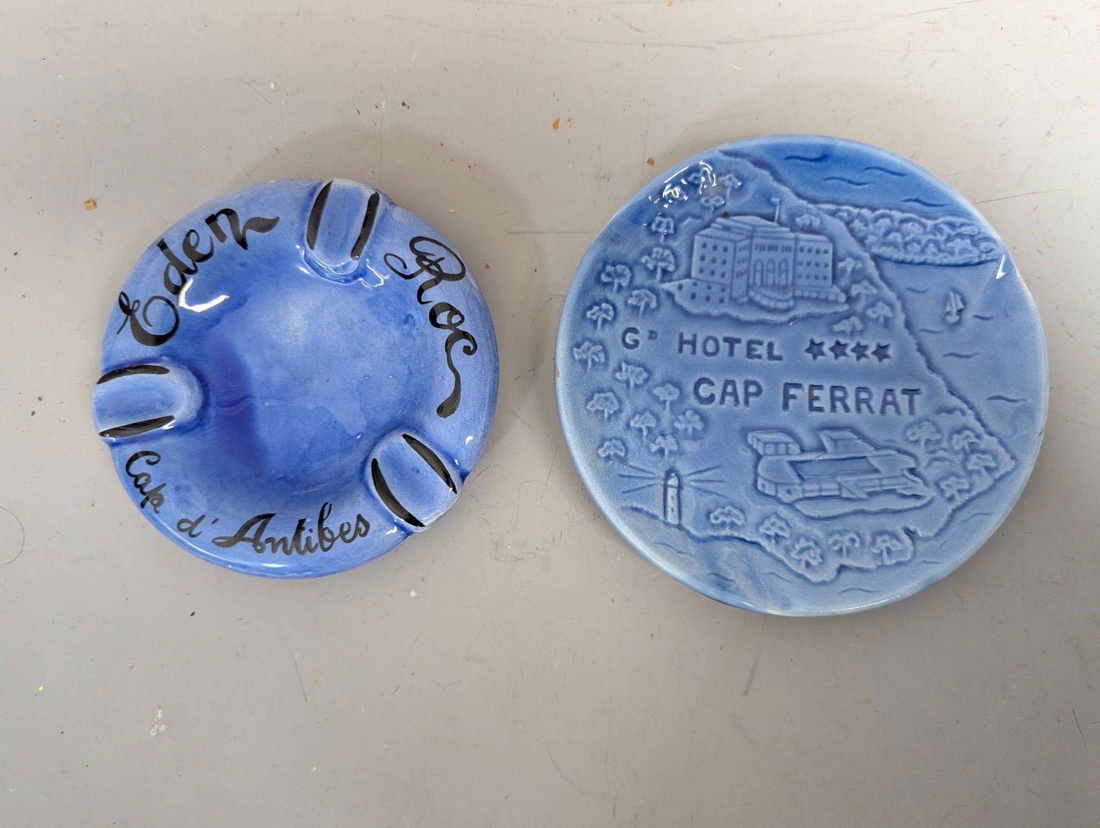 French Riviera Hotel Ceramic Ashtrays from 1960s: Two ceramic ashtrays from the French Riviera, dated to the 1960s. One ashtray, marked "Eden Roc Cap d'Antibes," features a deep blue glaze with black and white accents. The other ashtray depicts a det