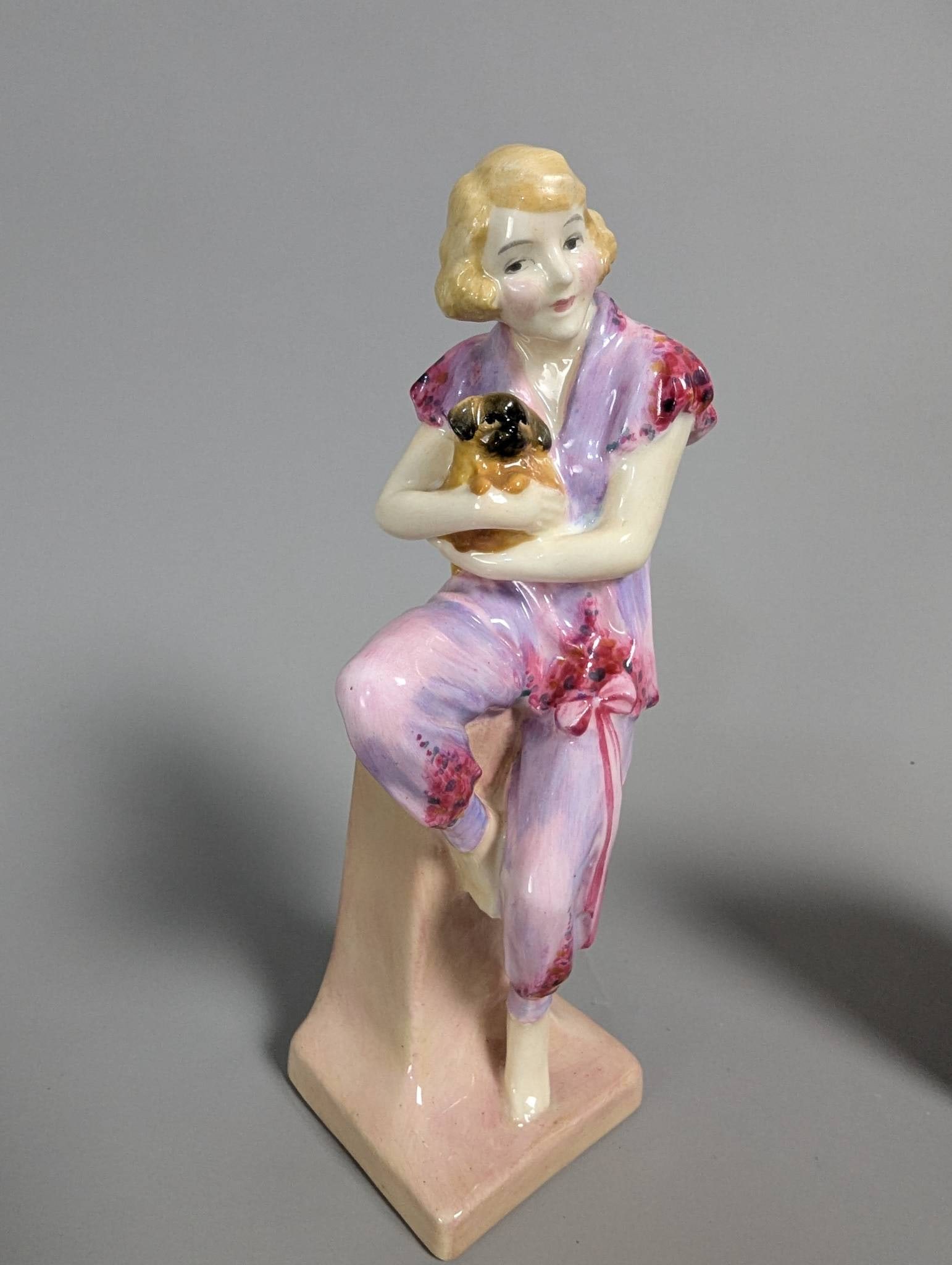 Royal Doulton HN1220 Lido Lady Figurine in Hand-Painted Attire: Royal Doulton HN1220 Lido Lady figurine, standing at 7 inches high. The piece features a seated woman dressed in delicate lavender and pink hand-painted attire, holding a small dog in her arms. Meticu