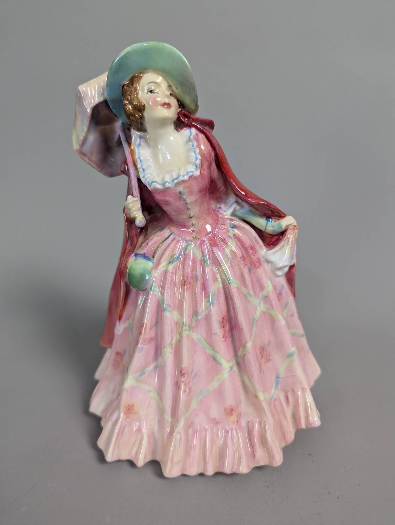 Royal Doulton HN 1744 Mirabel Figurine Depicting Elegant Woman: Royal Doulton porcelain figurine labeled HN 1744 Mirabel. Depicts an elegant woman in a vibrant pink dress with green and white accents, holding a parasol and wearing a red cape. Hand-painted with pre