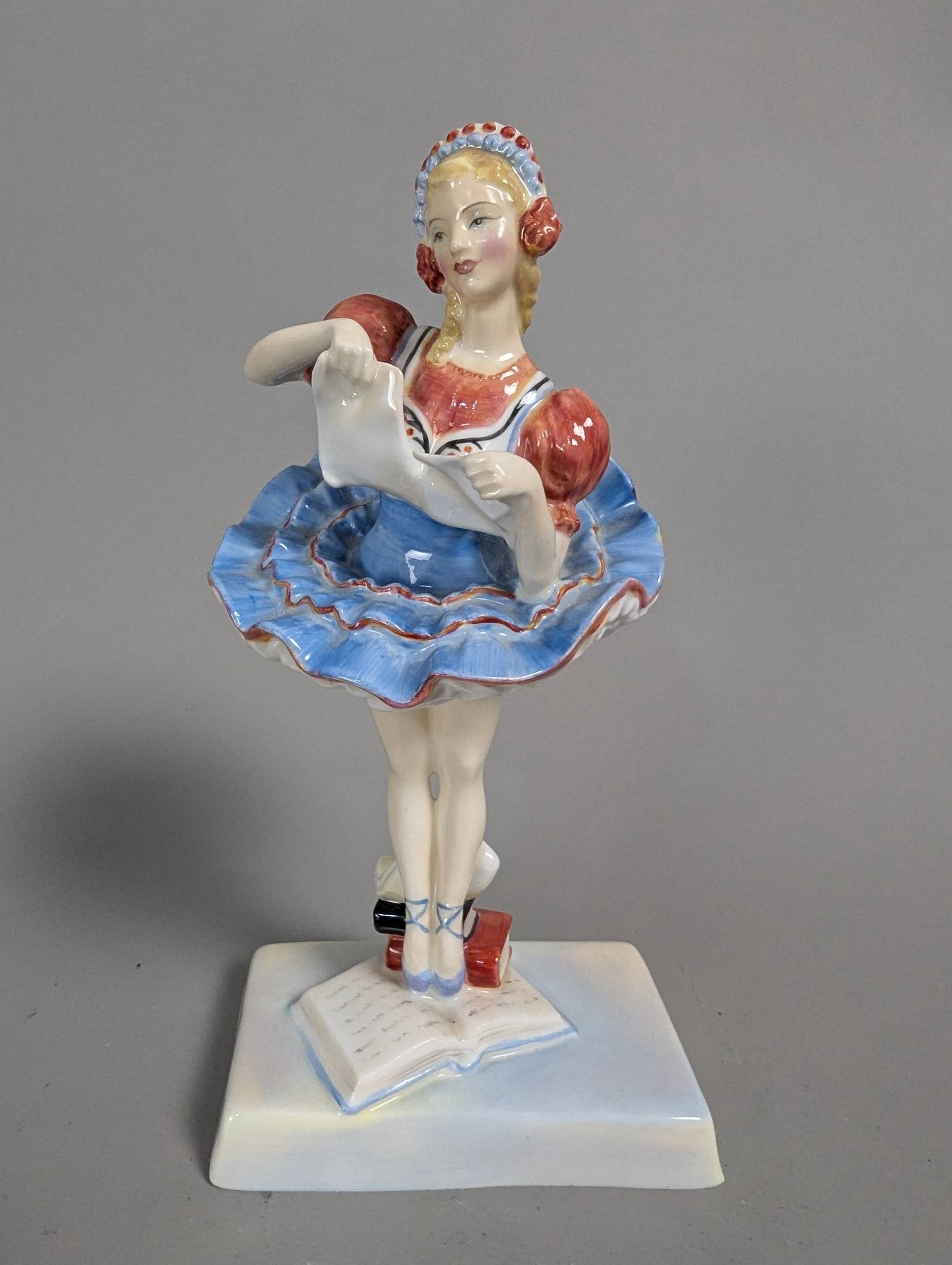 Royal Doulton HN 2115 Coppelia Porcelain Figurine: Royal Doulton Coppelia HN 2115 porcelain figurine, measuring 7 1/2 inches high. Modelled as a ballerina standing on one leg atop a base designed as a stack of books. The piece is painted in a vibrant