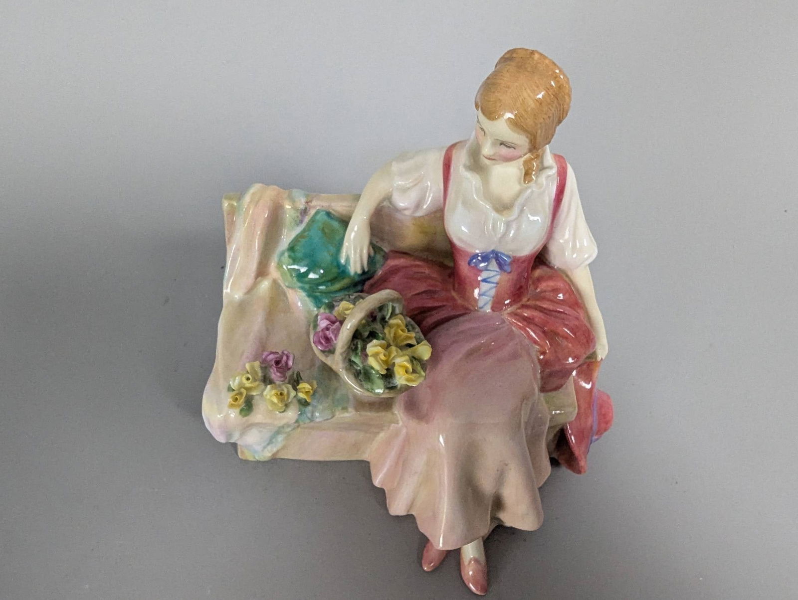 Royal Doulton HN 1899 Midsummer Noon Figurine: Royal Doulton HN 1899 figurine, titled Midsummer Noon. Crafted in fine bone china, this hand-painted decorative piece depicts a seated woman in a flowing dress holding a basket of flowers. Dimensions