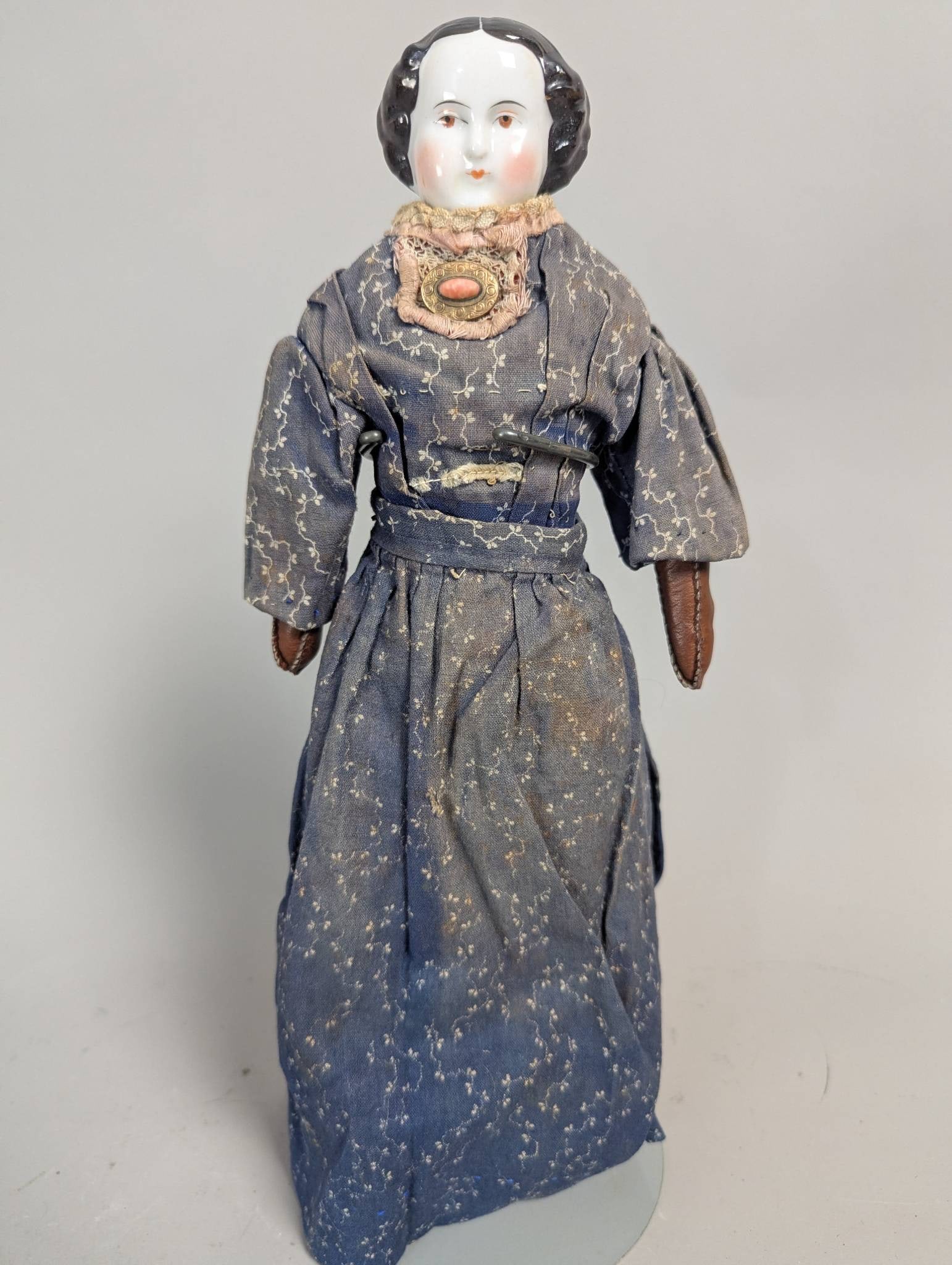 Antique Porcelain Doll with Black Hair and Aged Fabric Clothing: An antique porcelain doll, likely a parian or china doll from the mid-19th century, measuring 13 inches high. The doll features a distinct black hair design and rosy cheeks, with aged fabric clothing
